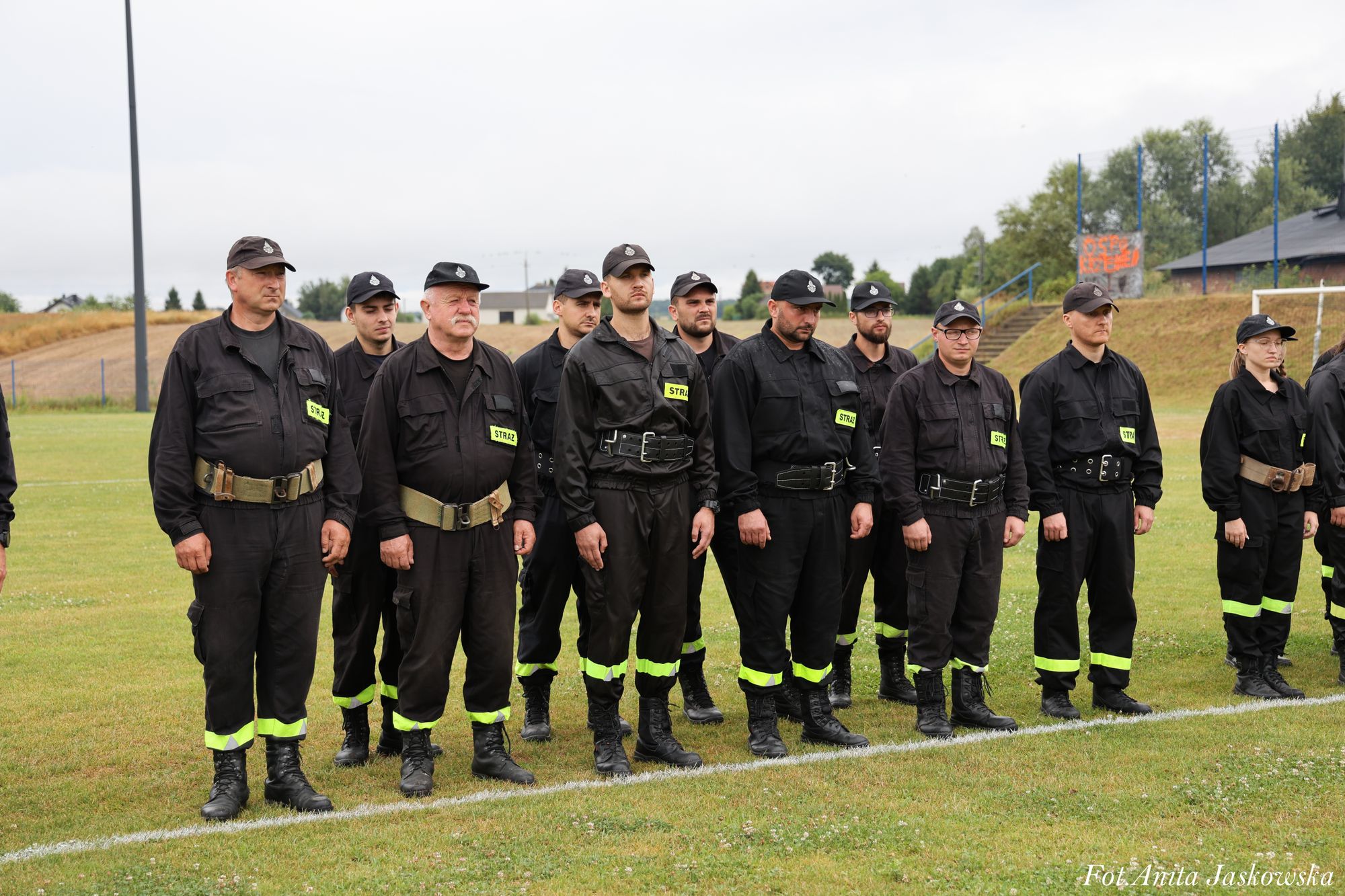 Grupa mężczyzn i kobiet w czarnych mundurach z żółtymi odblaskowymi paskami na nogawkach i czapkach, stojąca na zielonym trawniku na boisku sportowym.