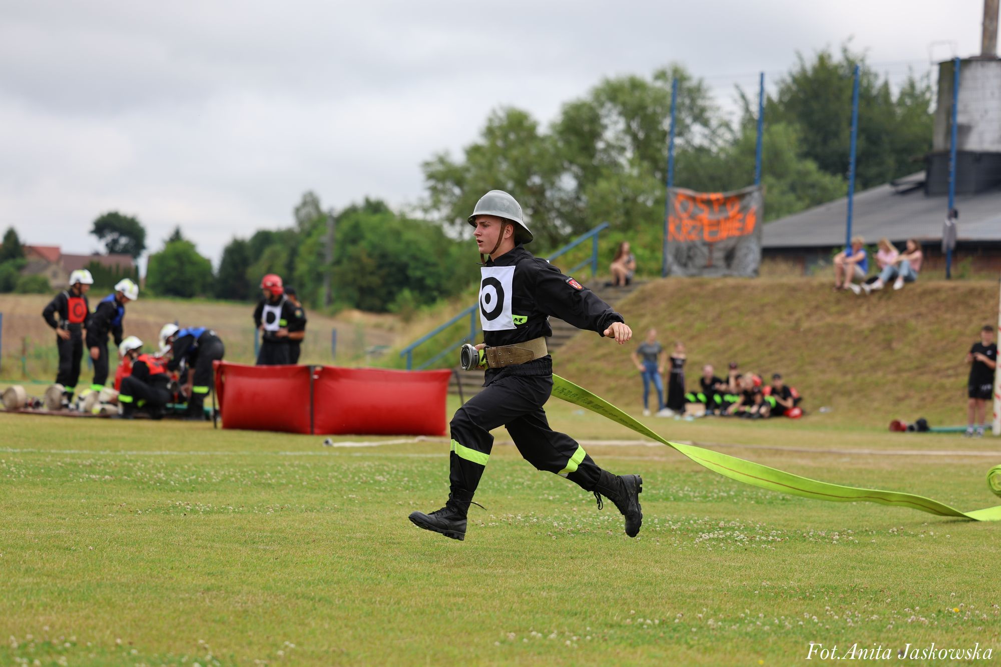 Mężczyzna w czarnym ubraniu strażackim, hełmie i z zielonym wężem strażackim biegnie po trawiastym boisku, w tle grupa osób i czerwone pojemniki.