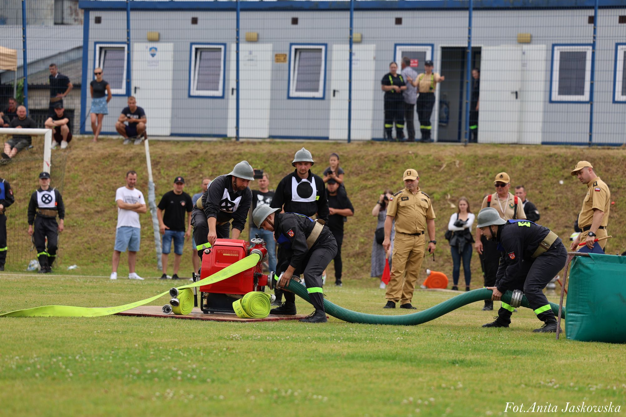 Strażacy w czarnych mundurach i hełmach podłączający zielony wąż do czerwonej pompy na zielonym boisku, w tle widoczni ludzie i budynki kontenerowe.