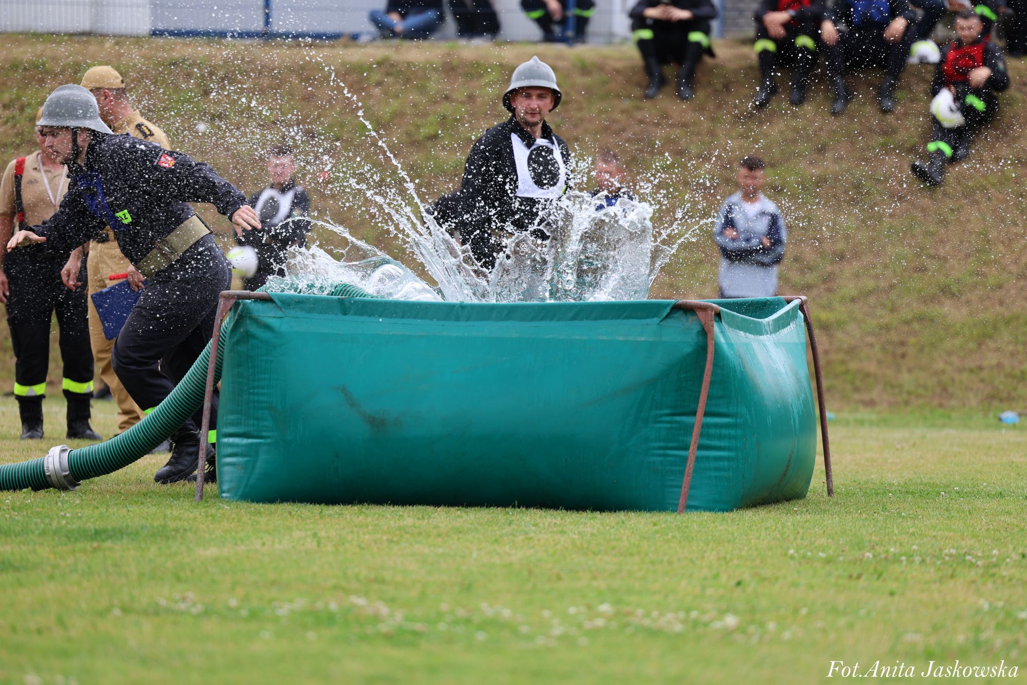 Dwóch mężczyzn w hełmach przy zielonym basenie z wodą, woda rozpryskuje się na zewnątrz, w tle trawa i osoby siedzące na wzniesieniu.
