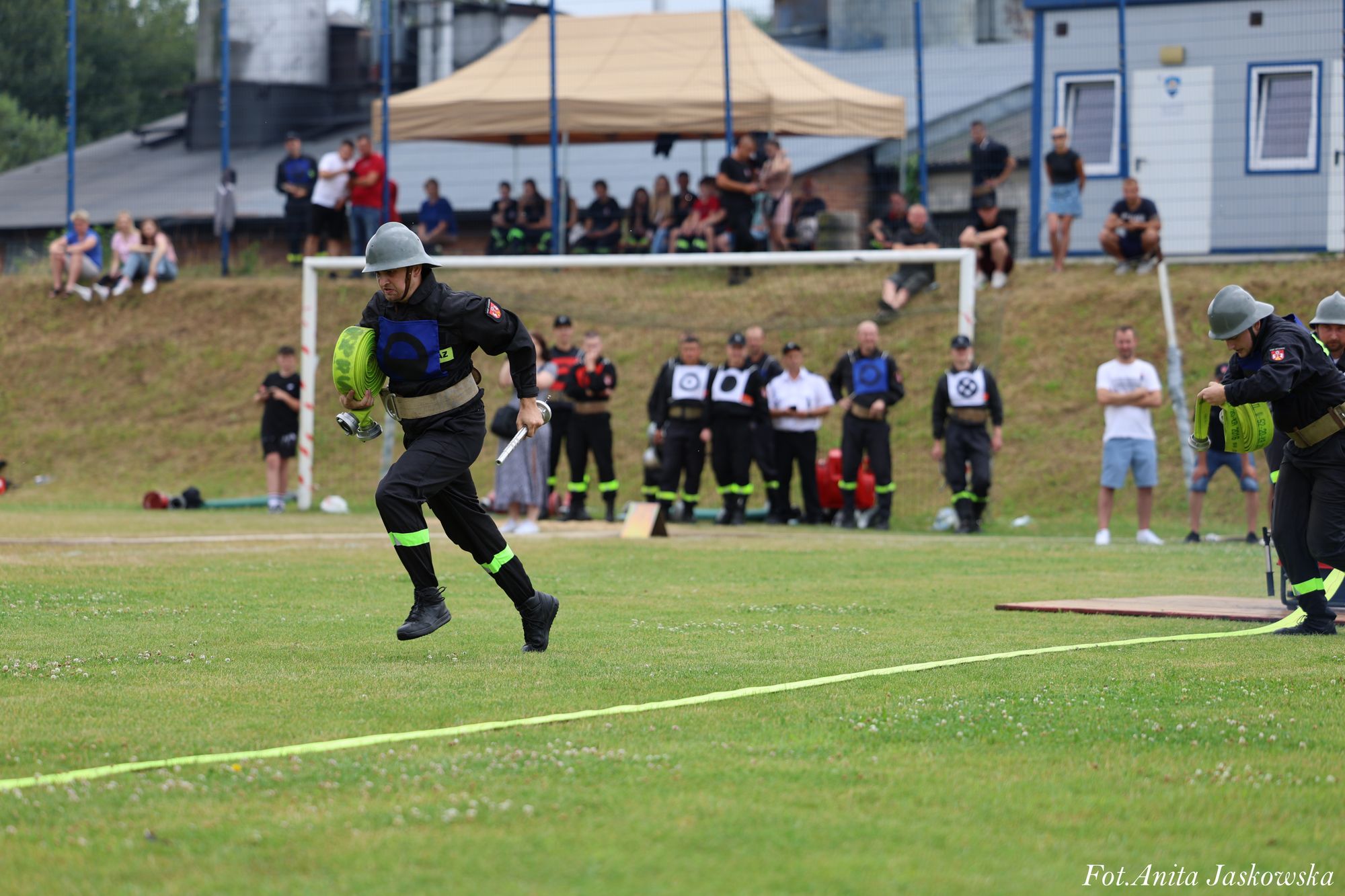 Strażacy w czarnych mundurach z odblaskowymi paskami i hełmach biegną po zielonej trawie, trzymając zwinięte zielone węże strażackie, w tle ludzie na skarpie i pod namiotem.