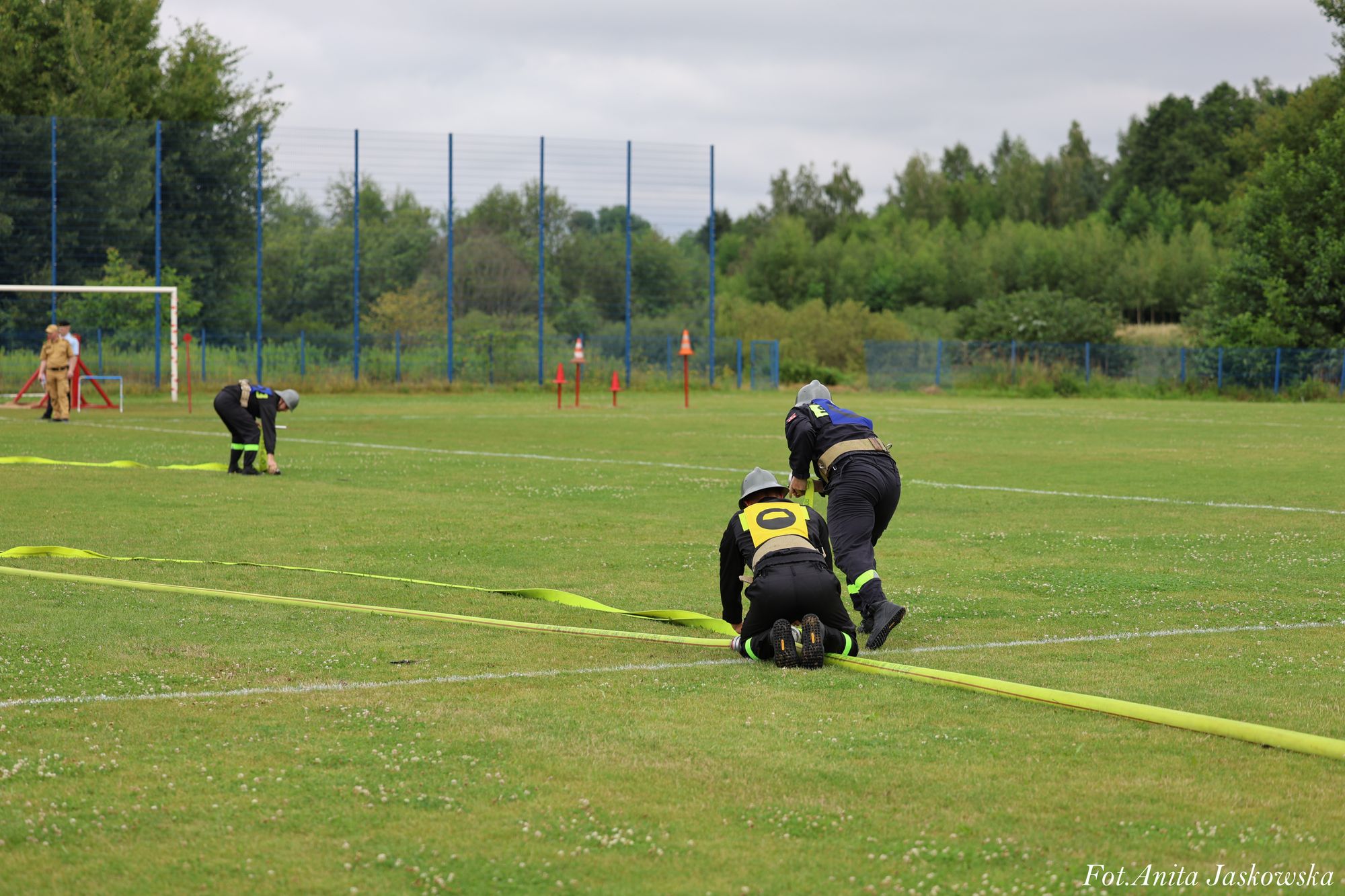 Dwoje strażaków w czarnych mundurach i hełmach rozkłada żółty wąż na zielonym boisku, w tle drzewa i ogrodzenie.