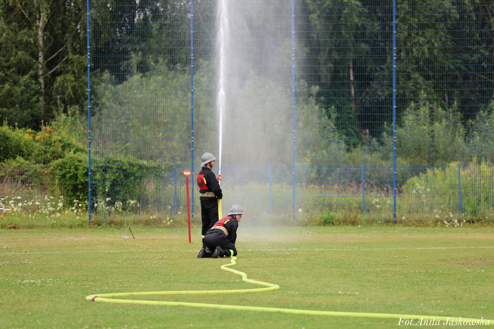 Dwaj strażacy w czarnych uniformach i hełmach trzymają żółty wąż gaśniczy, z którego leci strumień wody, na zielonym trawniku z siatką i drzewami w tle.