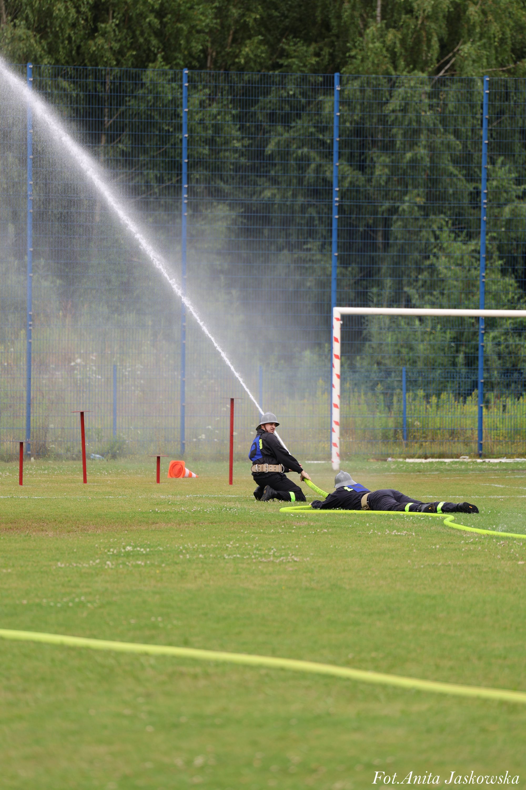 Dwóch strażaków w kaskach na zielonym boisku, jeden klęczy, drugi leży, trzymają żółty wąż z którego leci silny strumień wody, w tle niebieska siatka i bramka.