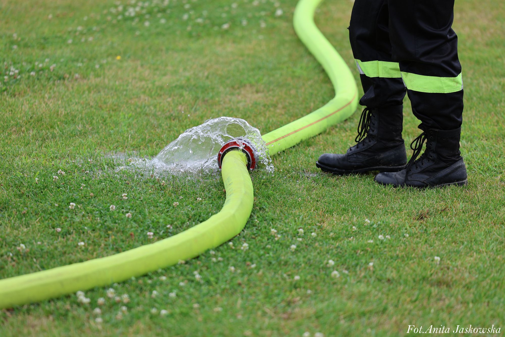 Czarna odzież robocza z odblaskowymi pasami, czarne buty stojące na trawie obok zielonego węża strażackiego z wyciekającą wodą.