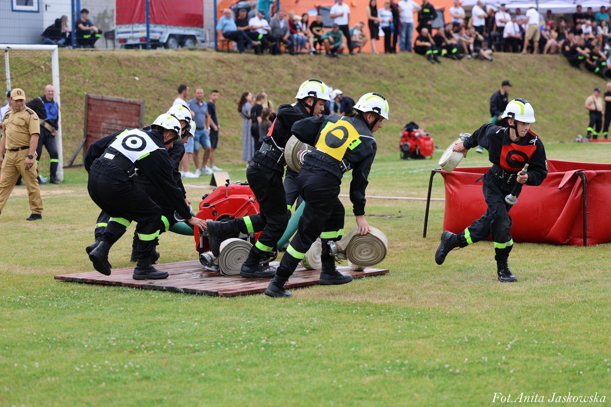 Grupa strażaków w czarnych uniformach z odblaskowymi paskami i białych hełmach rozwija zwinięte węże na zielonym boisku, w tle widoczni kibice i sędzia.