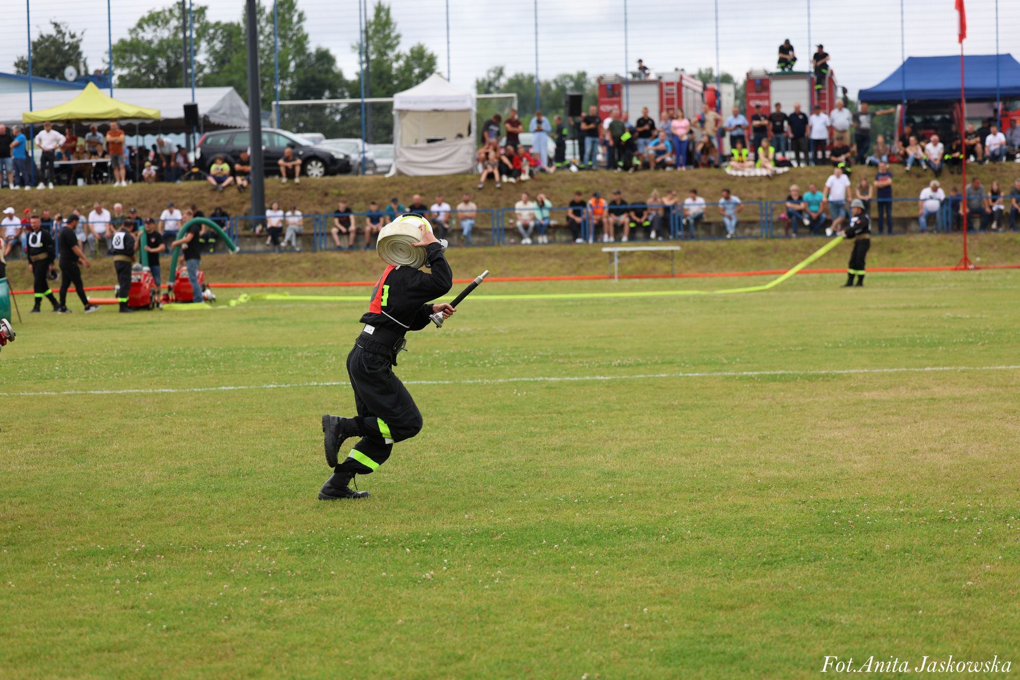 Strażak w czarnym mundurze z neonowymi elementami biegnie po zielonym boisku, trzymając zwinięty biały wąż strażacki na ramieniu, w tle widownia i sprzęt ratowniczy.