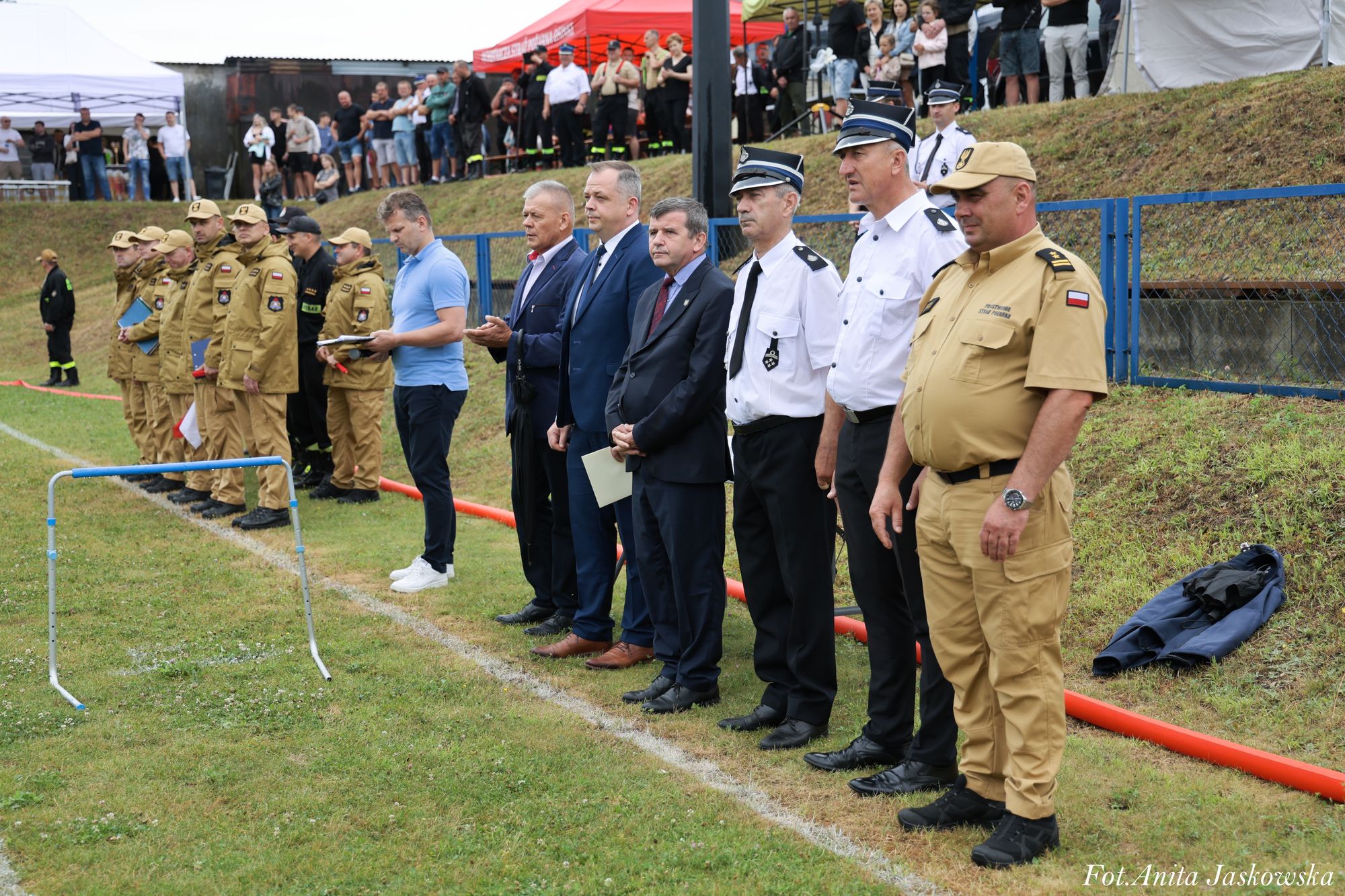Grupa mężczyzn w mundurach i garniturach stojąca na trawie, w tle ludzie na wzniesieniu i namioty, niebieskie ogrodzenie, czerwony wąż strażacki.