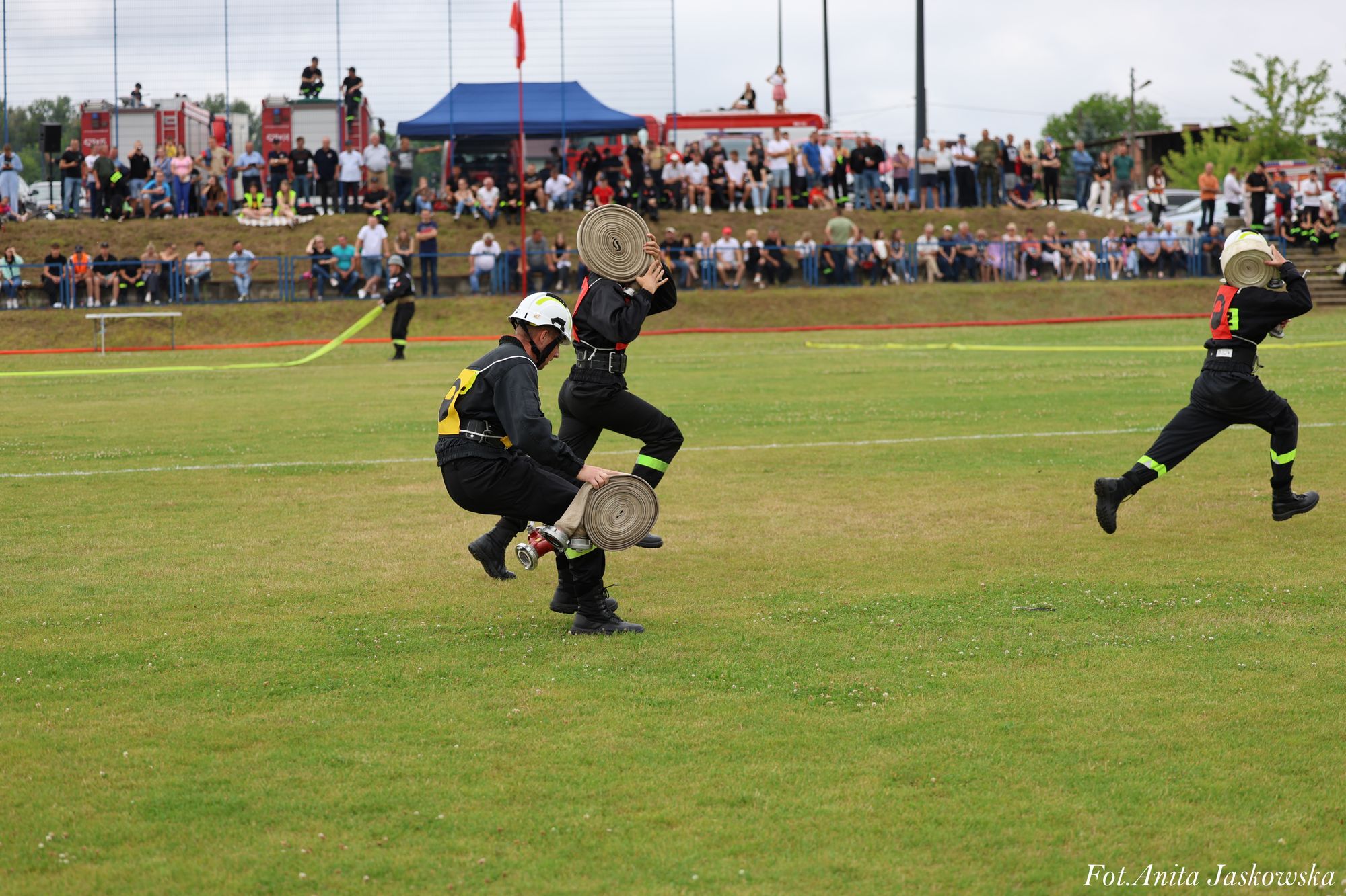 Trzech strażaków w czarnych mundurach i białych hełmach biegnie po zielonym boisku, każdy niesie zwinięty wąż strażacki, w tle tłum ludzi i wozy strażackie.