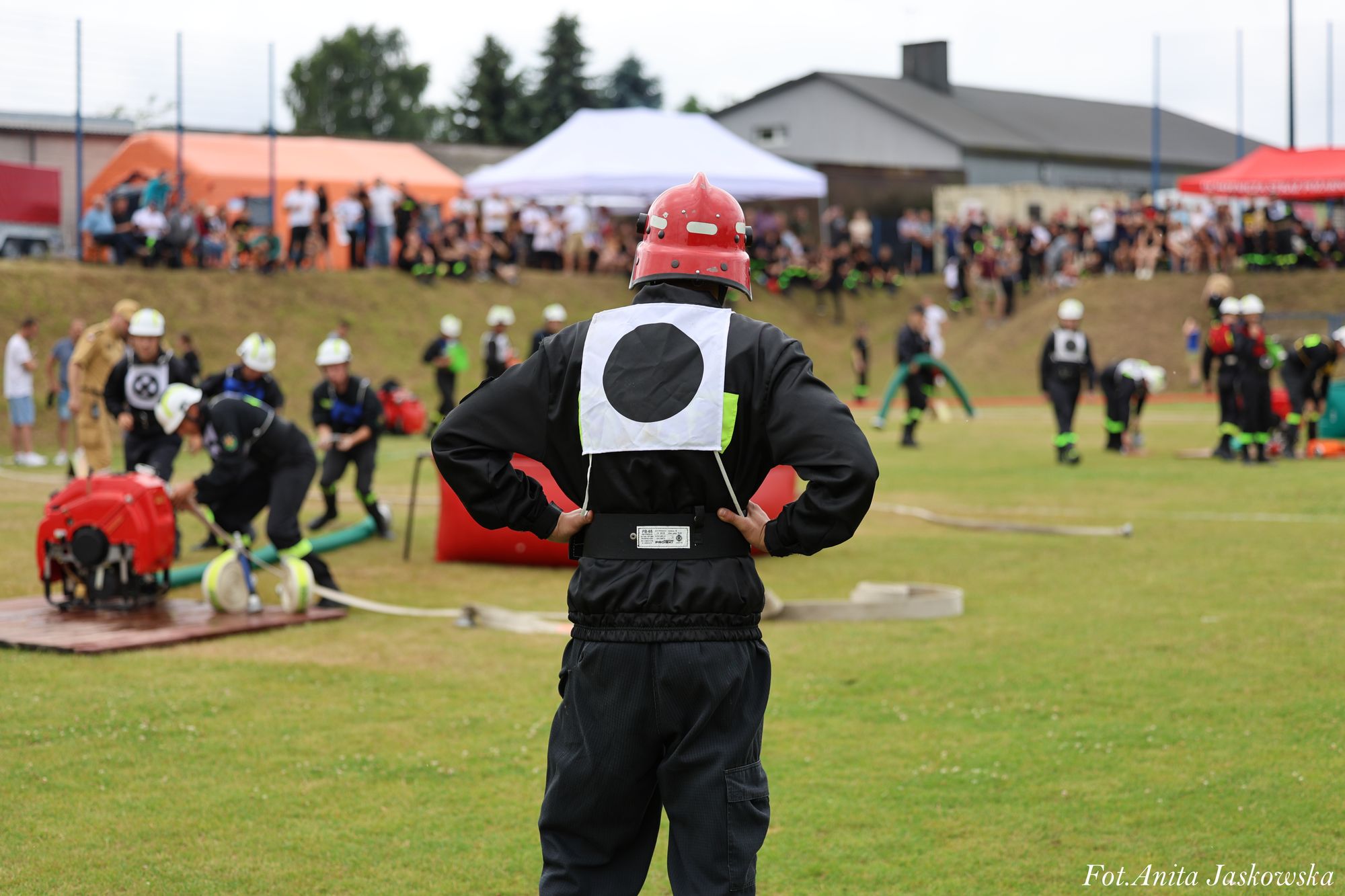 Strażak w czarnym ubraniu i czerwonym hełmie z białą plakietką z czarnym kołem na plecach stoi na zielonym trawniku, w tle inne osoby i namioty.