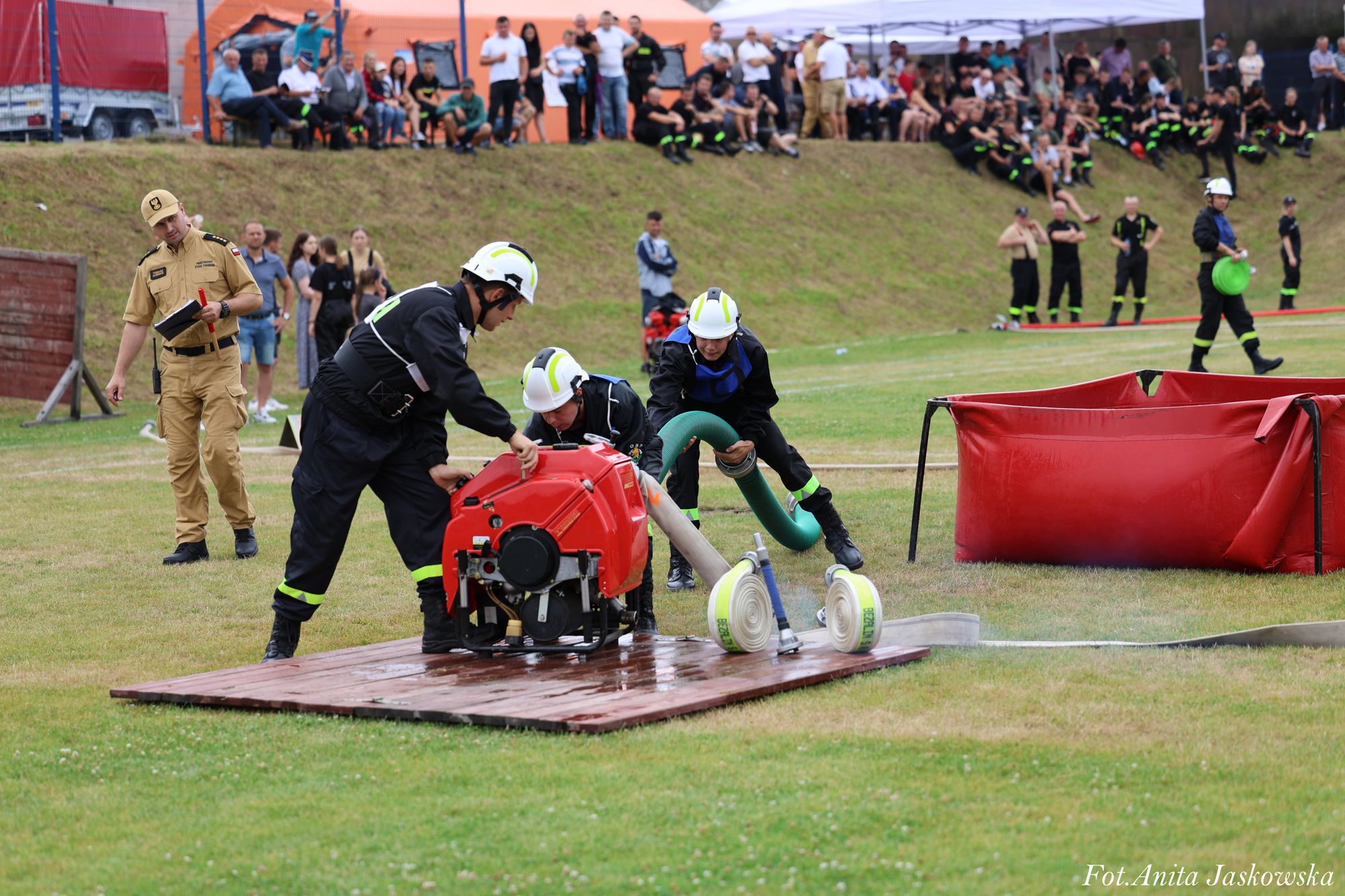 Trzech strażaków w czarnych mundurach i białych hełmach obsługuje czerwoną pompę na drewnianej platformie, obok zwinięte białe węże i czerwony zbiornik na trawie, w tle widzowie.