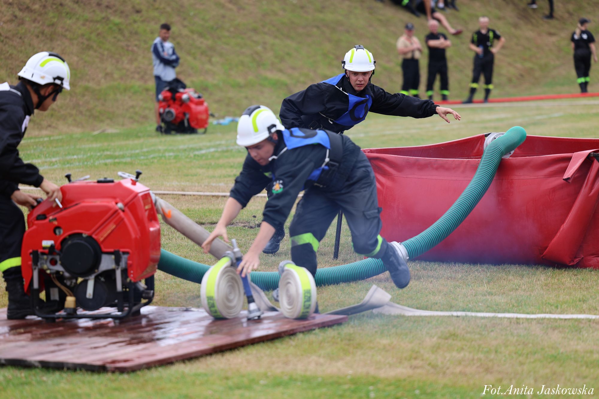 Trzech strażaków w kaskach i niebieskich kamizelkach przy czerwonym sprzęcie pożarniczym na trawie, zielony wąż podłączony do czerwonego zbiornika.