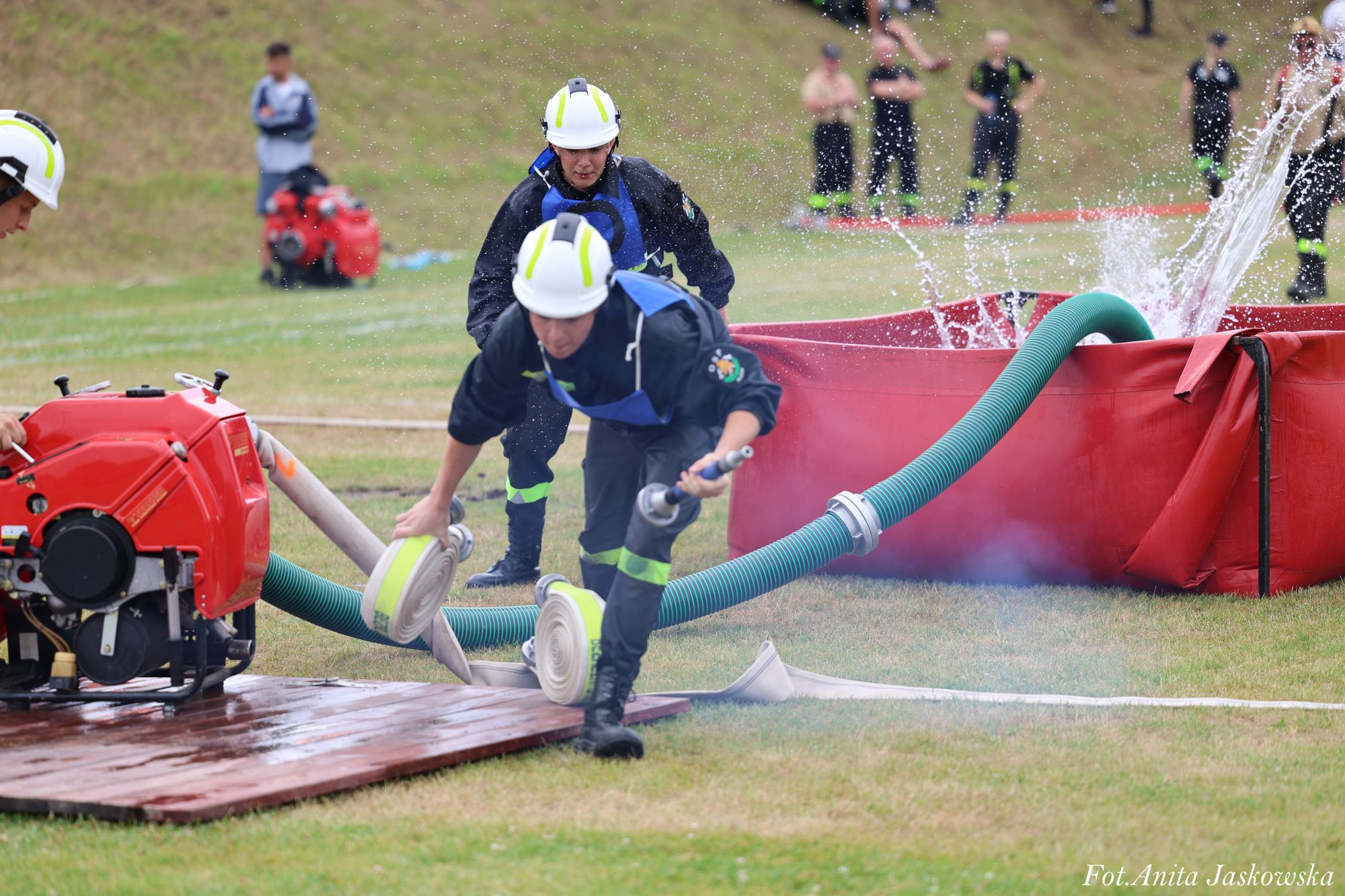 Trzech strażaków w kaskach i granatowych uniformach rozwija białe węże połączone z czerwoną przenośną cysterną i pompą na trawie.