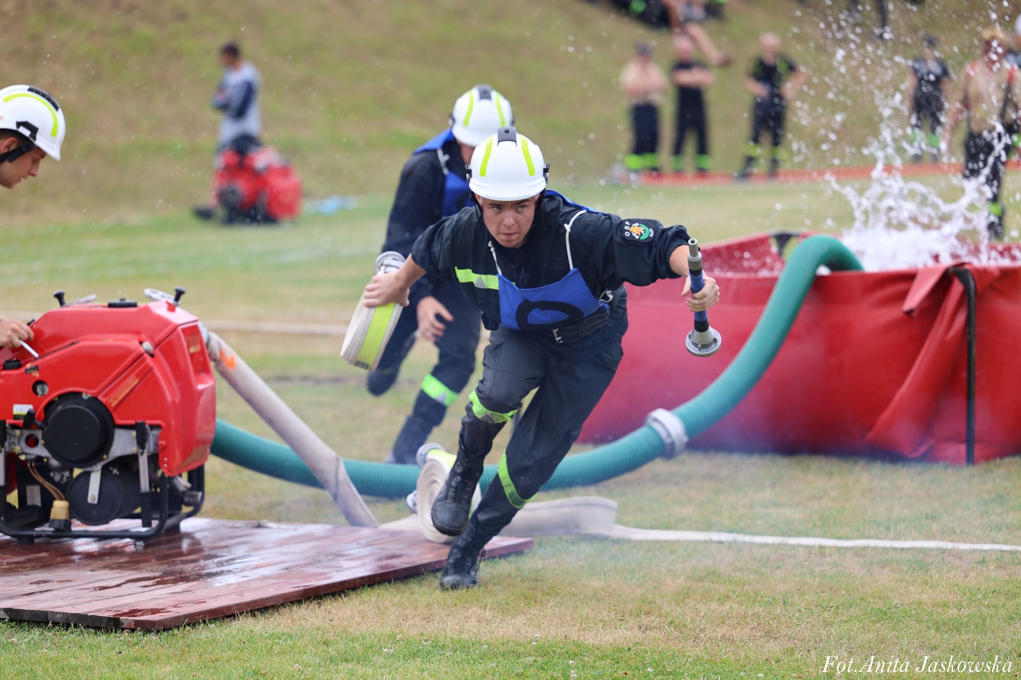 Mężczyzna w kasku i czarnym uniformie z niebieskim fartuchem biegnący z wężem strażackim na zielonej trawie, rozpryski wody, w tle czerwona pompa i ludzie.