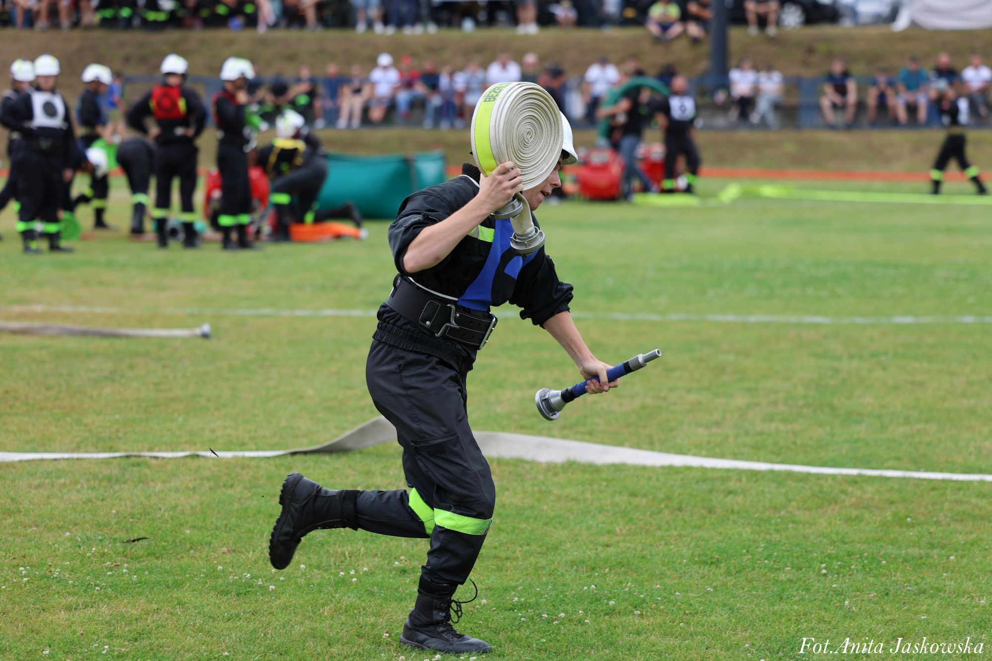 Strażak w czarnym uniformie z neonowymi wstawkami i białym hełmem biegnie po zielonym boisku, trzymając zwinięty biały wąż strażacki i metalową końcówkę.
