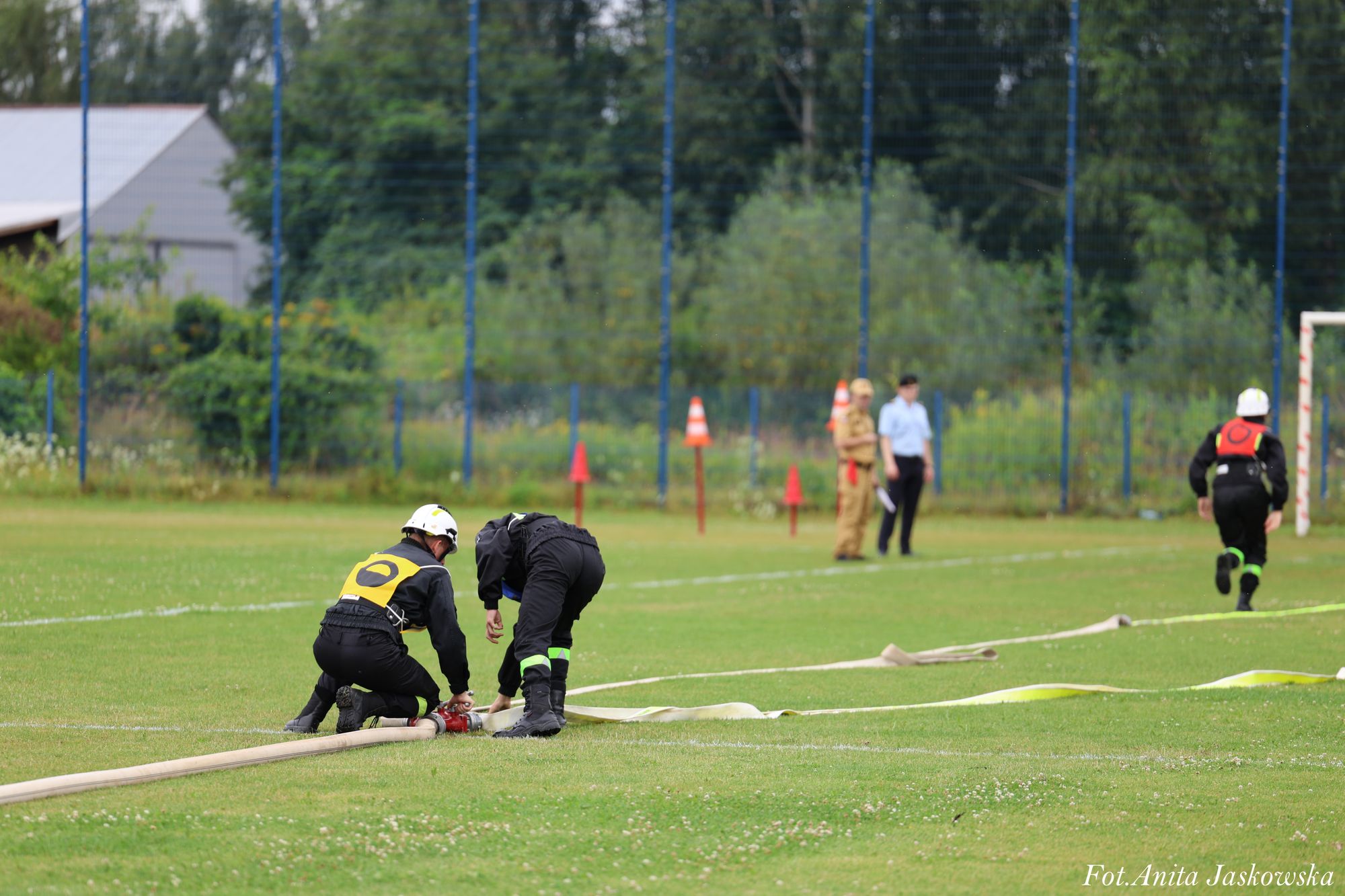 Dwie osoby w czarnych uniformach i białych hełmach rozkładają wąż strażacki na trawniku, trzecia biegnie, w tle dwie osoby obserwują, zieleń i siatka ogrodzeniowa.