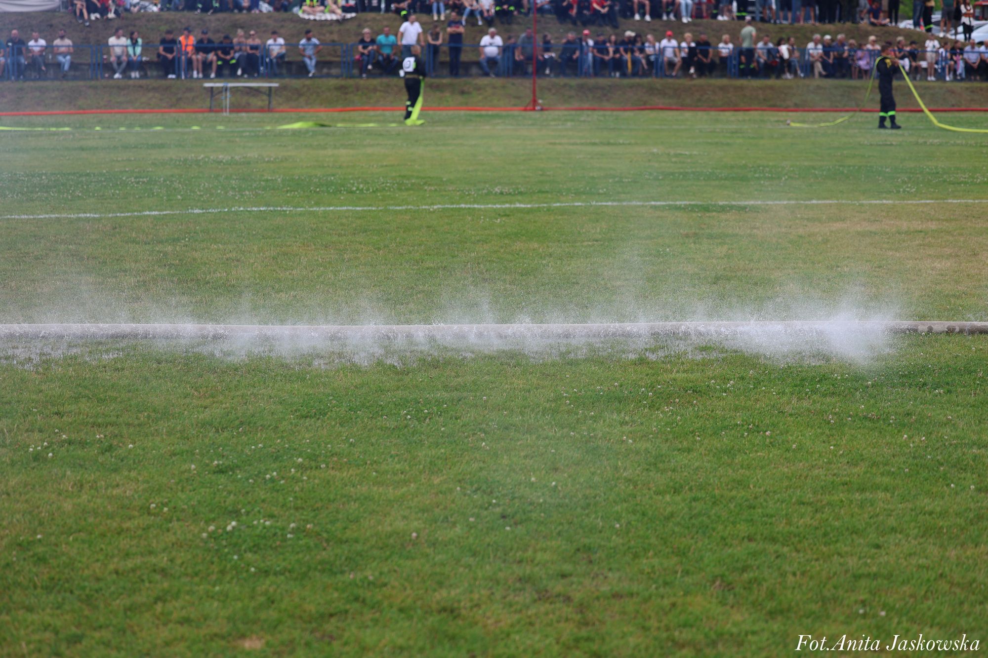 Zielona trawa boiska, na niej rozproszona mgiełka wody, w tle ludzie siedzący na murawie i dwóch mężczyzn w czarnych ubraniach z neonowymi elementami trzymających żółte węże.