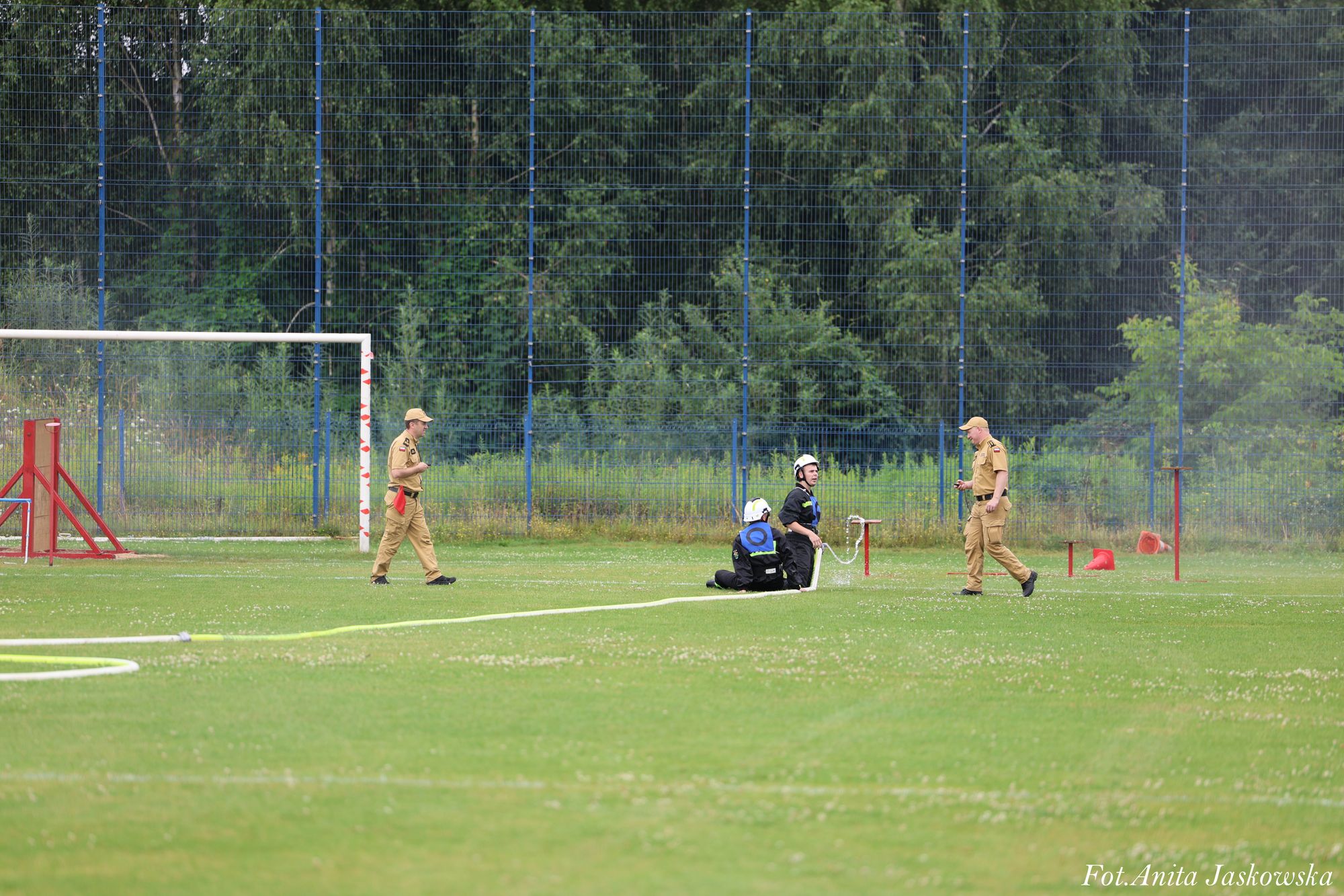 Trzech mężczyzn na zielonym boisku, dwóch w czarnych ubraniach z hełmami, jeden w beżowym mundurze, w tle wysoka niebieska siatka i drzewa.