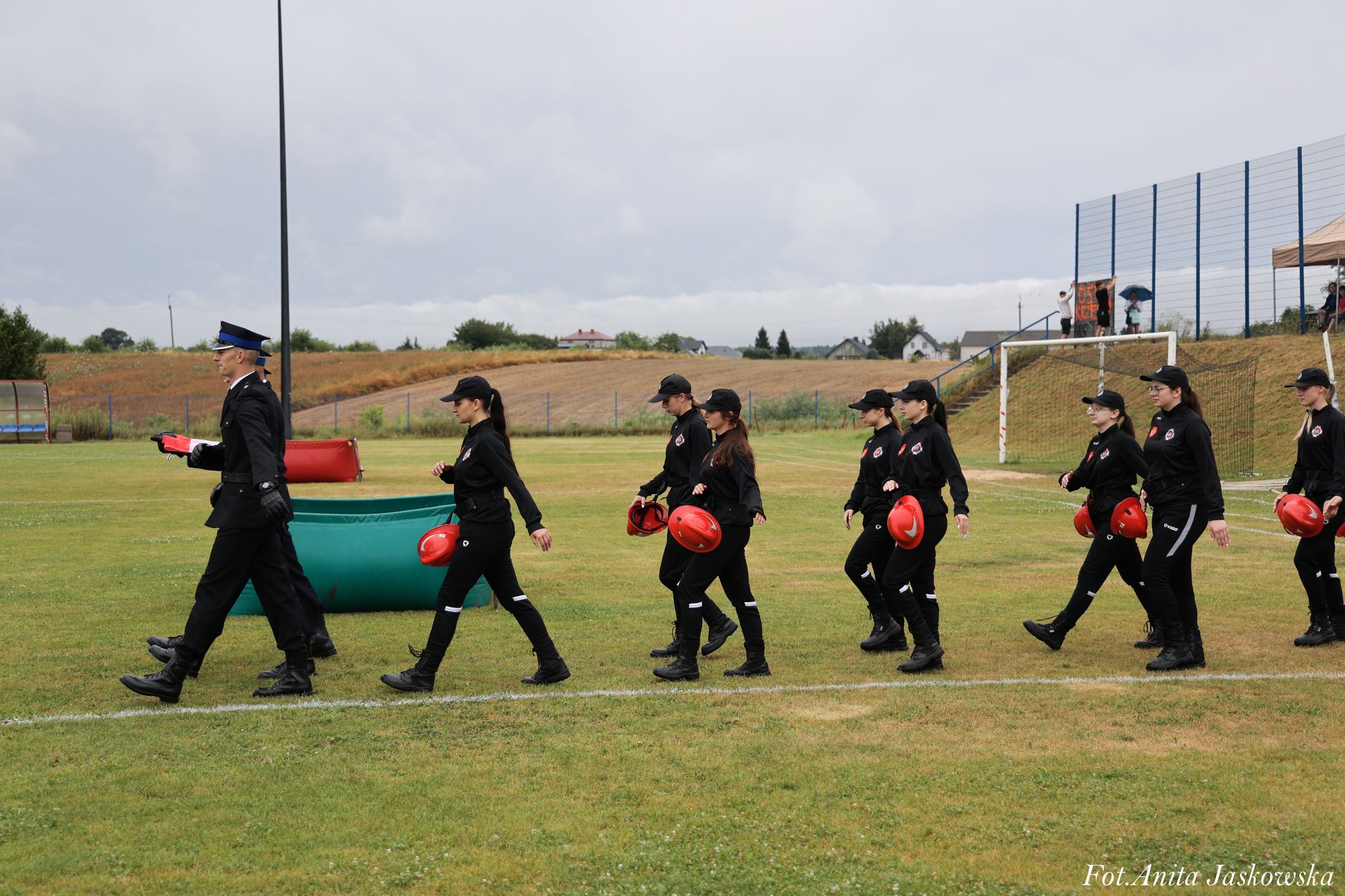Grupa osób w czarnych mundurach i czapkach maszeruje po trawie, niektóre trzymają czerwone hełmy, w tle boisko sportowe i wiejskie pola.