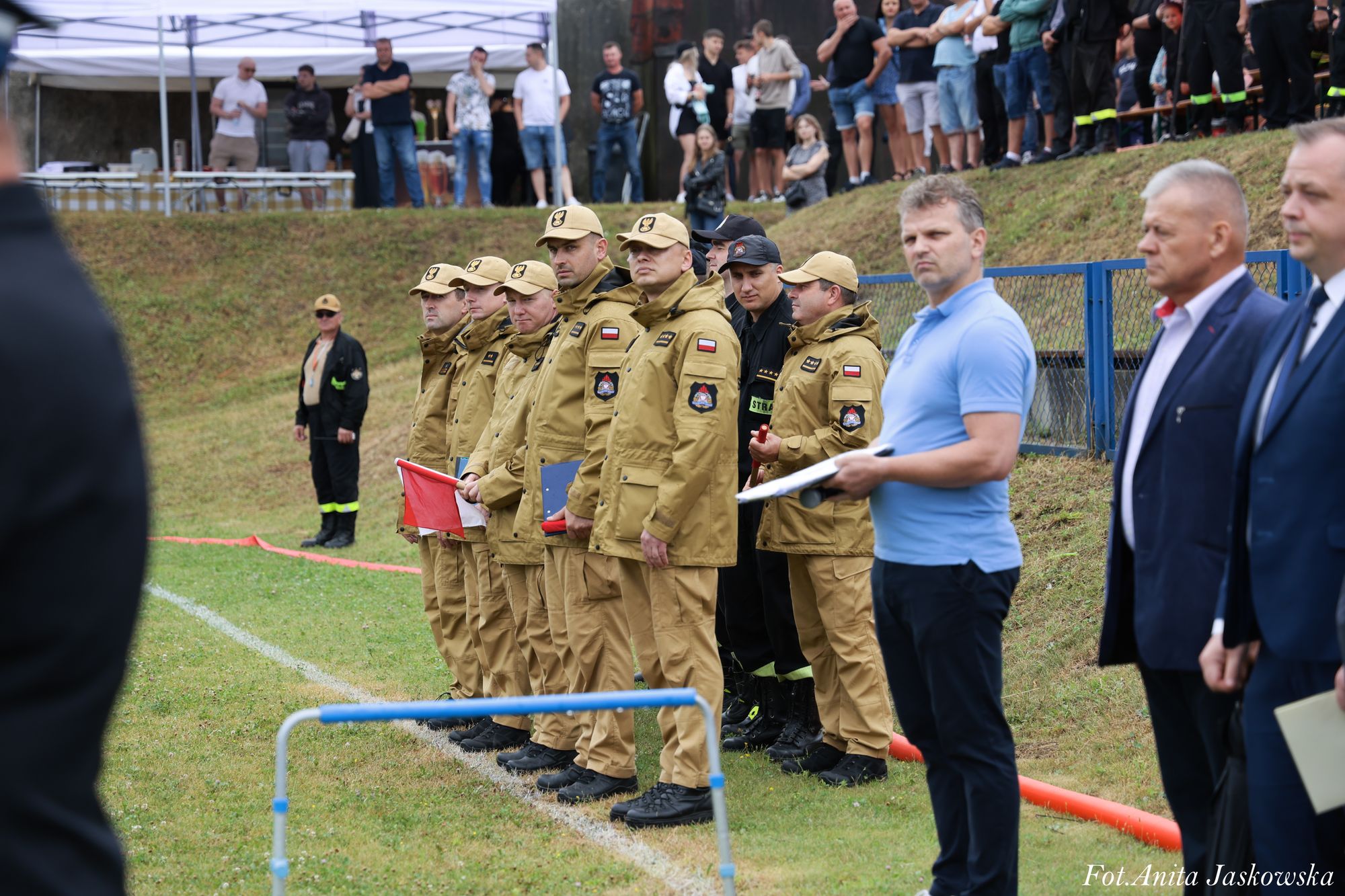 Grupa mężczyzn w mundurach khaki i kilku w cywilnych ubraniach stoi na trawie przed skarpą z ludźmi, w tle biały namiot i ogrodzenie niebieskie.