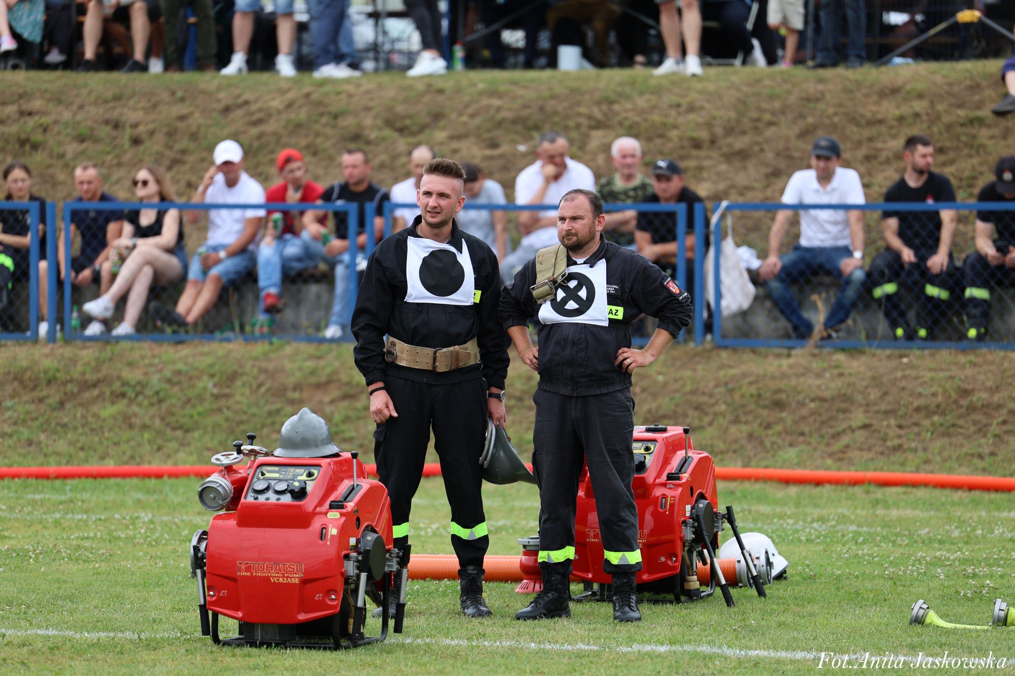 Dwóch mężczyzn w czarnych mundurach strażackich stoi na zielonej trawie obok dwóch czerwonych pomp pożarniczych, w tle niebieska barierka i siedzący ludzie.