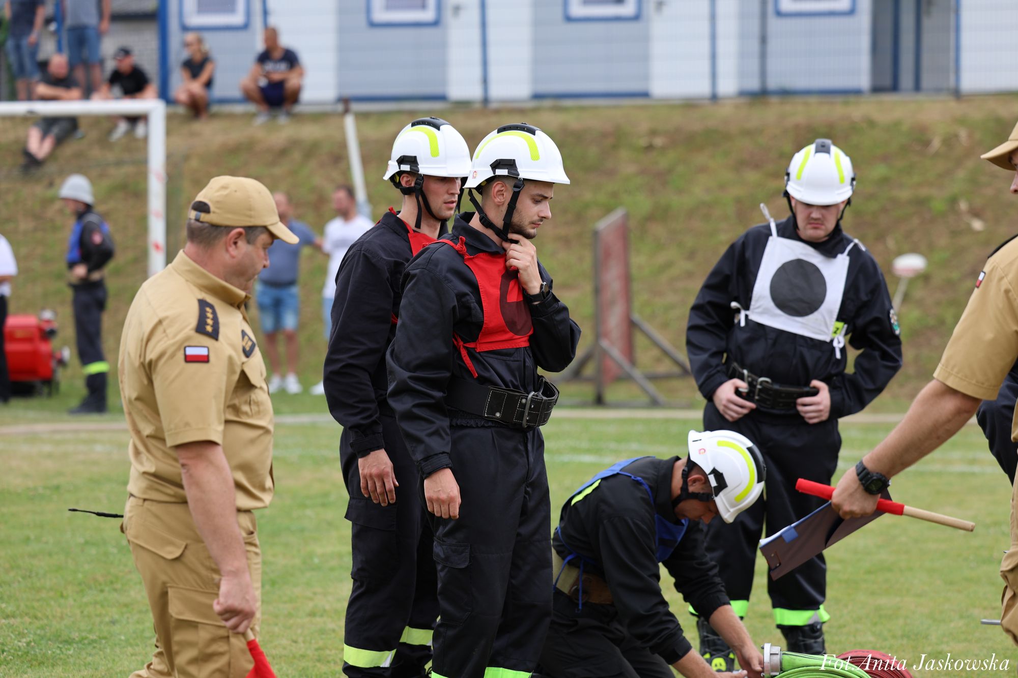 Cztery osoby w czarnych uniformach i białych hełmach, dwie z czerwonymi kamizelkami, jedna w niebieskiej, jedna z okrągłym czarnym znakiem na białym tle, dwie osoby w beżowych mundurach na zielonej trawie.