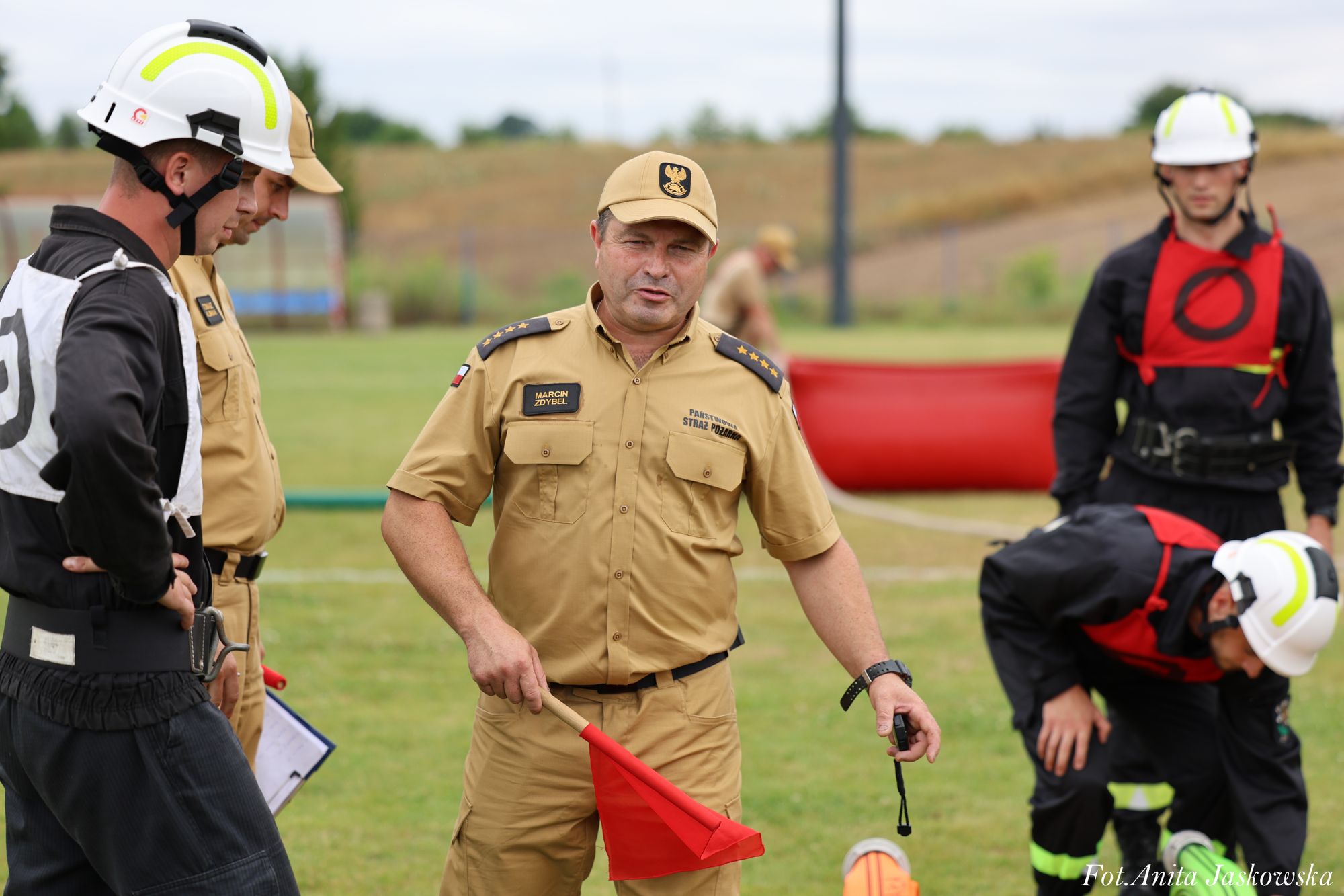 Mężczyzna w beżowym mundurze z naszywkami i czapką trzyma czerwony trójkątny sygnał i stoper, wokół niego inni mężczyźni w hełmach i czarnych mundurach z czerwonymi kamizelkami.