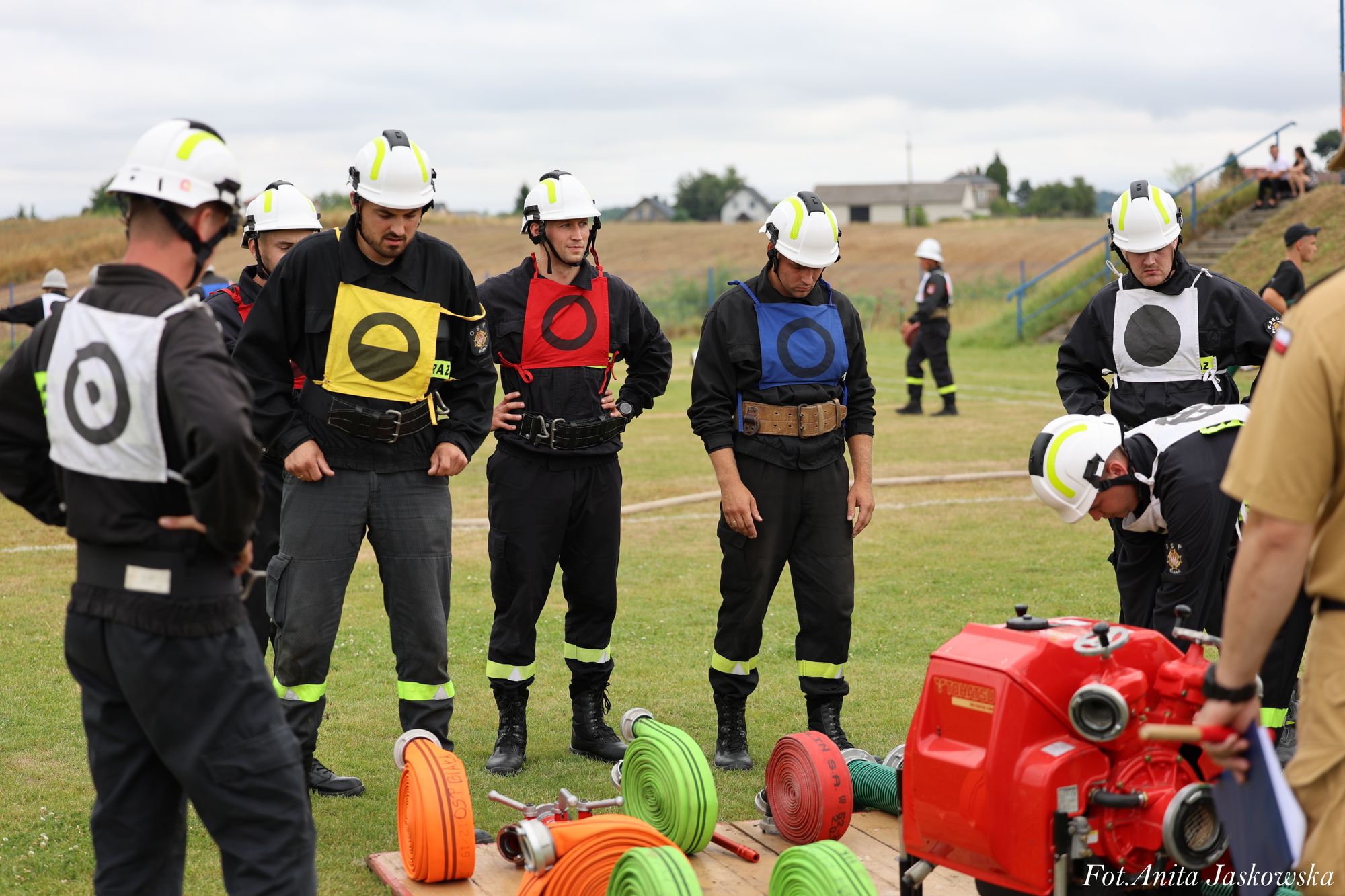 Grupa mężczyzn w czarnych mundurach i białych hełmach stoi na trawie wokół zwiniętych węży strażackich w kolorach pomarańczowym, zielonym i czerwonym oraz czerwonej pompy wodnej.