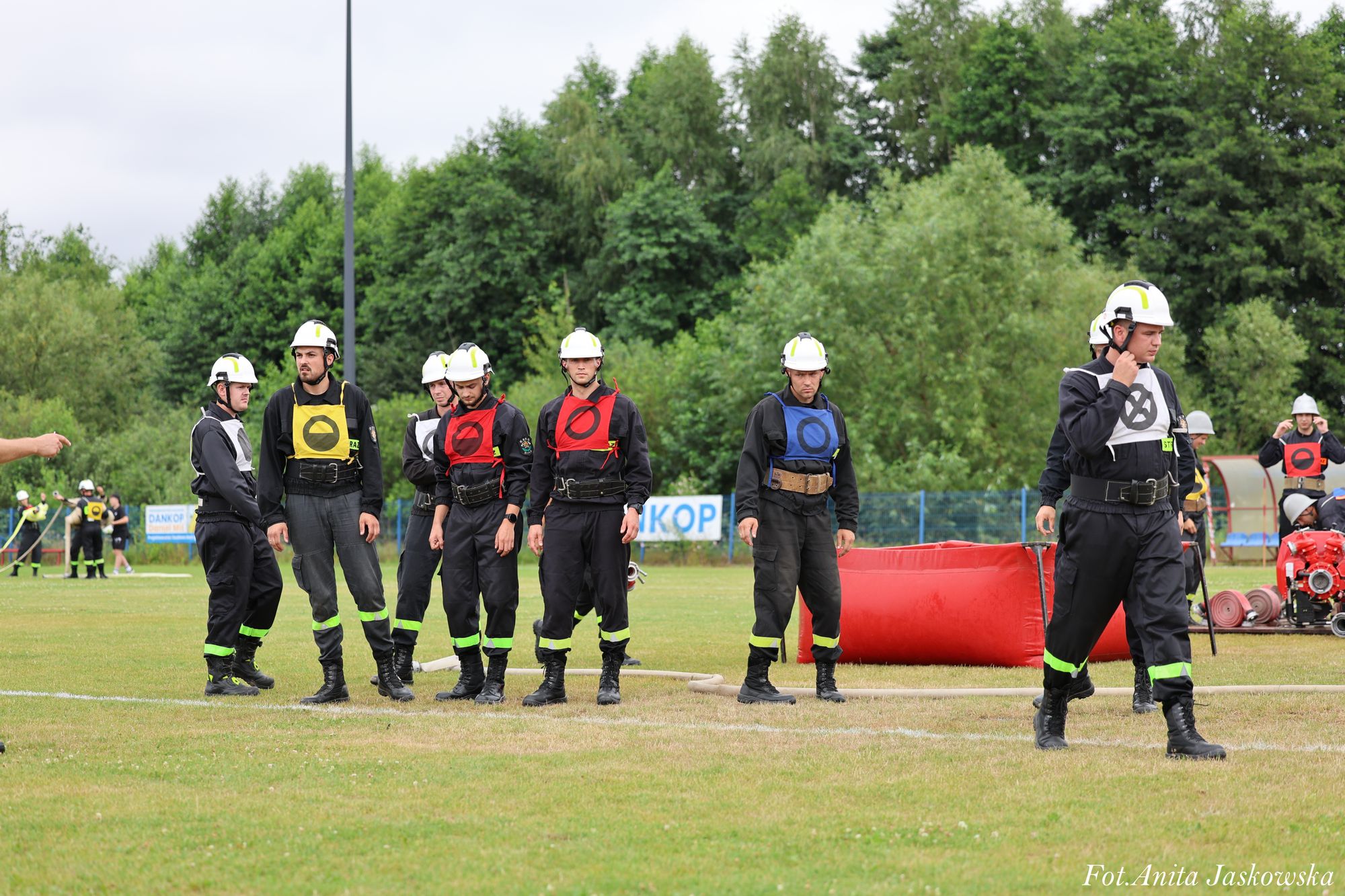 Grupa ludzi w mundurach strażackich z kolorowymi oznaczeniami na kamizelkach i białych kaskach stoi na trawiastym boisku, w tle drzewa i sprzęt strażacki.