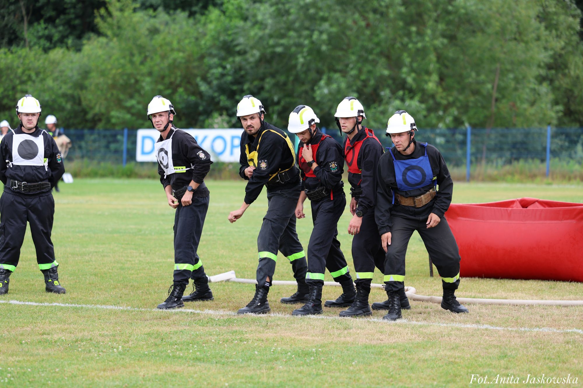 Sześciu mężczyzn w kaskach i ubraniach strażackich o różnych kolorach oznaczeń na trawie, za nimi czerwona dmuchana konstrukcja i zielone drzewa.