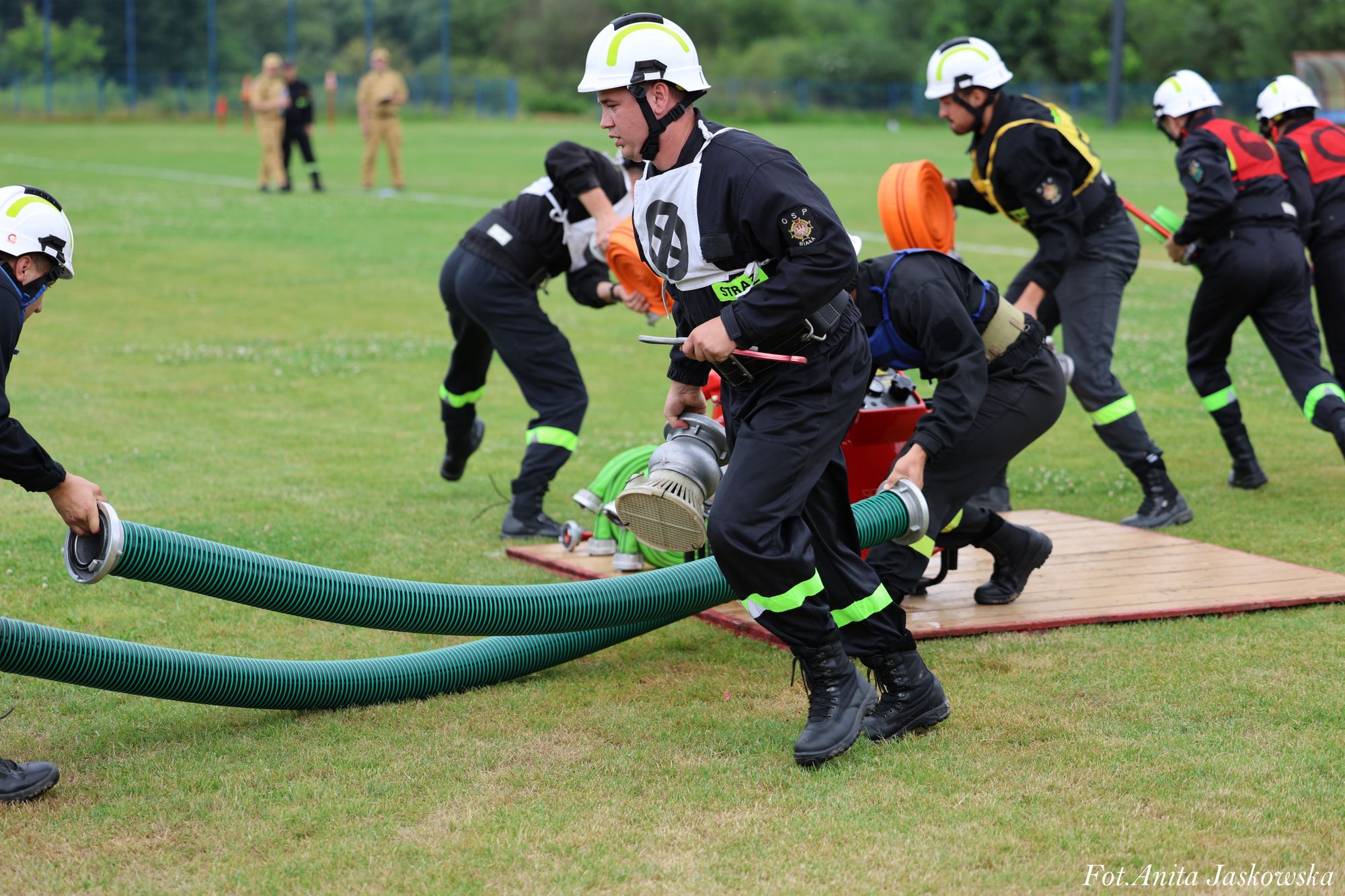 Grupa strażaków w czarnych mundurach i białych hełmach na zielonym trawniku rozkłada zielone i pomarańczowe węże pożarnicze podczas ćwiczeń.