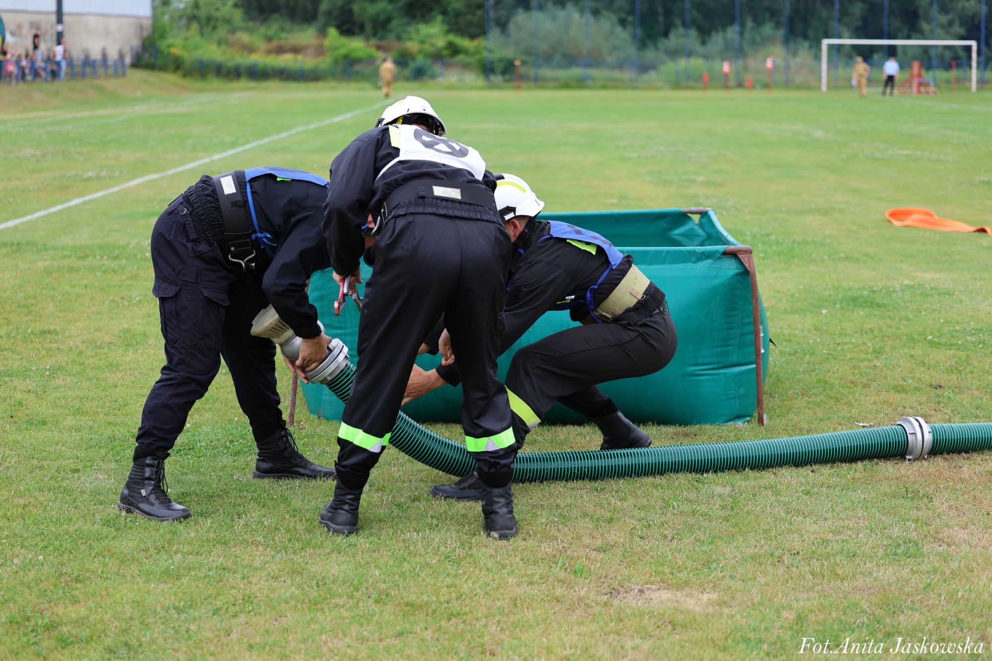 Trzy osoby w mundurach i kaskach na zielonym trawiastym boisku montują zielony elastyczny wąż do metalowej ramy z zielonym płótnem.