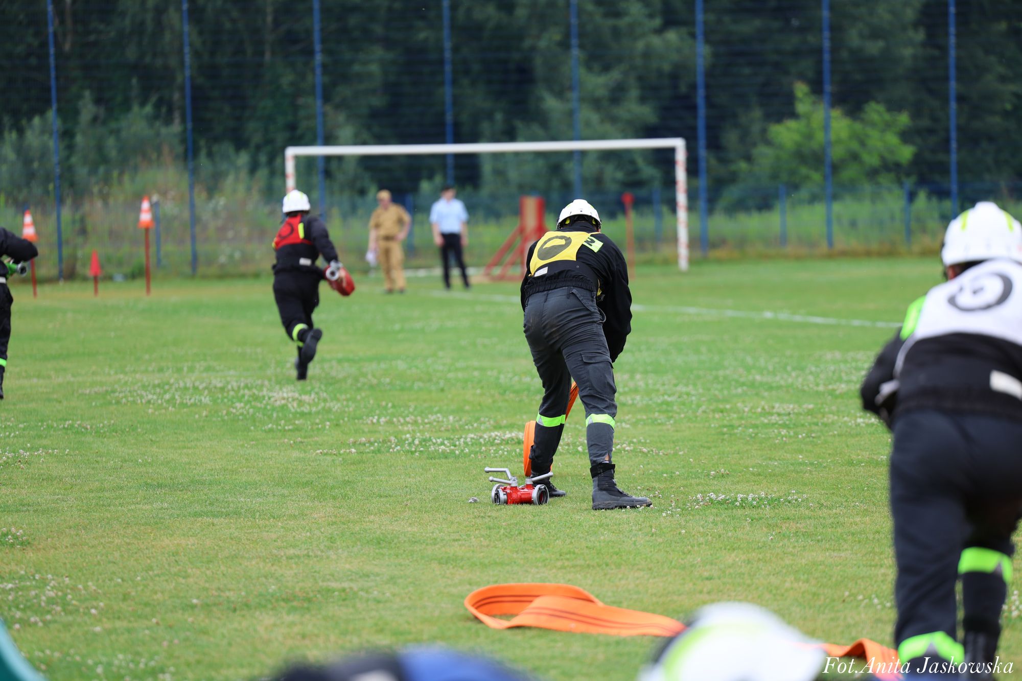 Trzech osób w ciemnych kombinezonach i kaskach na zielonym boisku, jedna z nich obsługuje czerwony sprzęt, w tle bramka i zalesnione otoczenie.