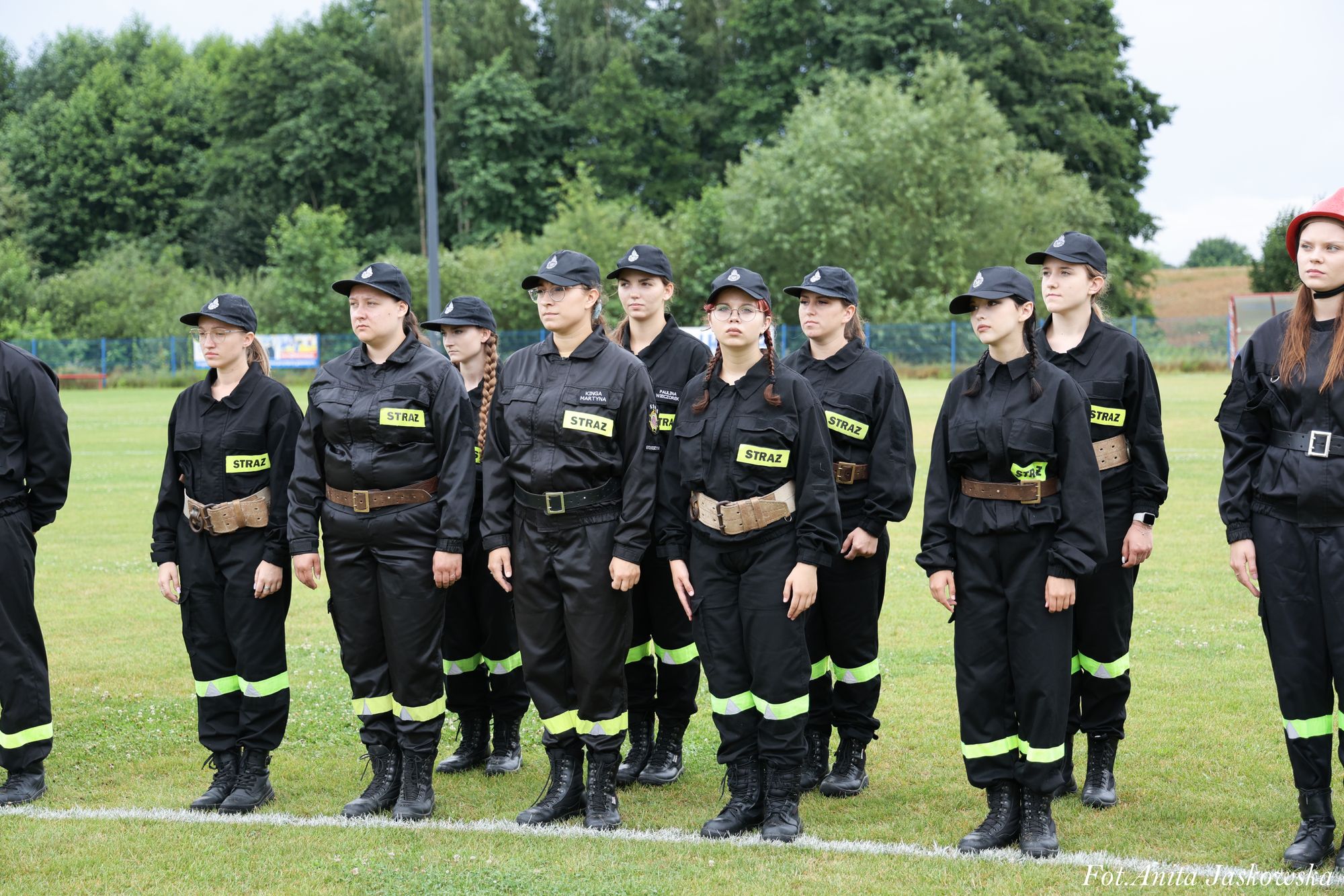 Grupa kobiet w czarnych mundurach z napisem "STRAŻ" na tle zielonej trawy i drzew. Niektóre mają czapki, jedna czerwoną hełm.