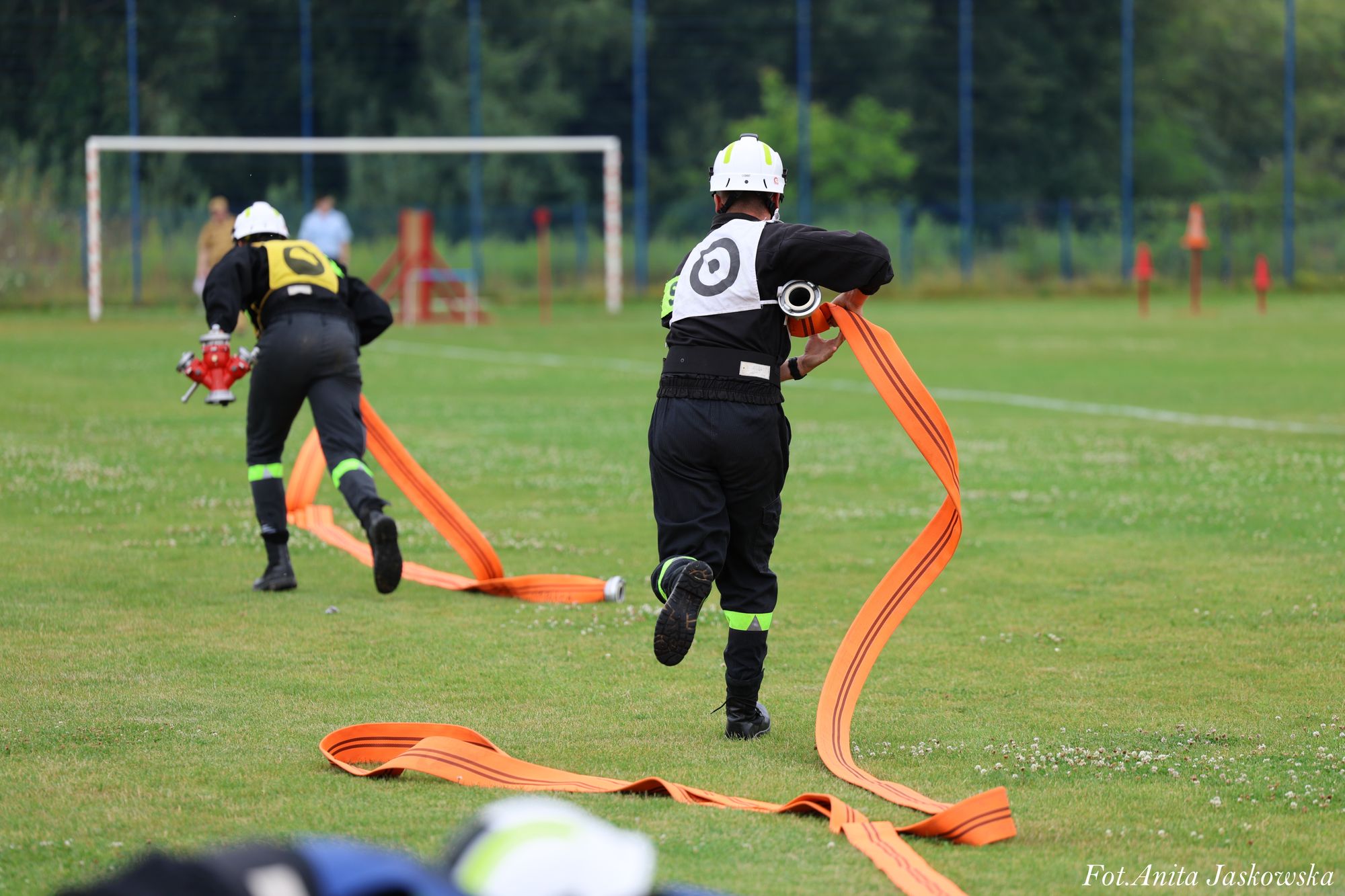 Dwóch strażaków w czarnych mundurach i białych hełmach biegnie po trawiastym boisku, niosąc rozwinięte pomarańczowe węże pożarnicze.