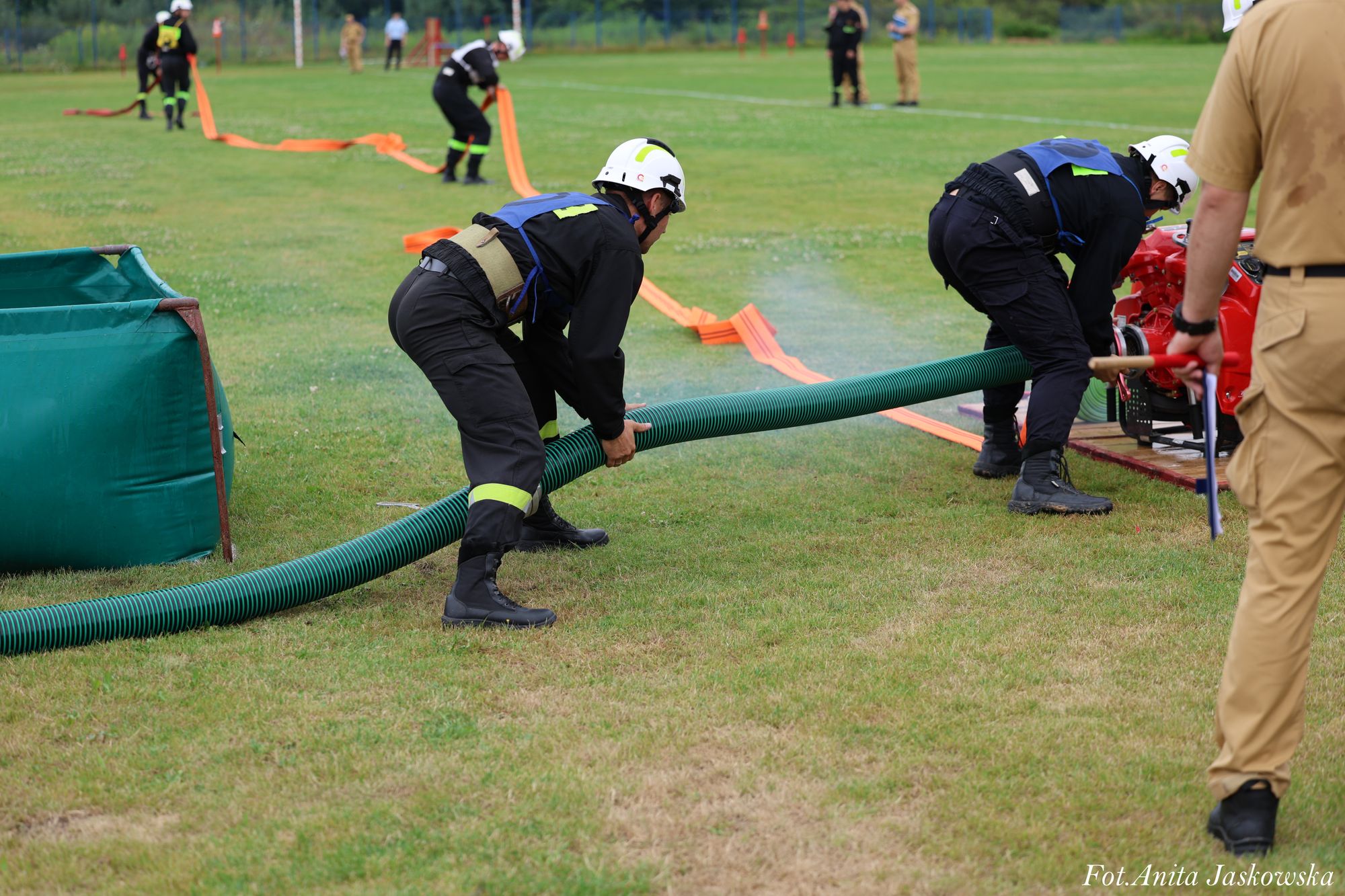 Dwóch strażaków w czarnych mundurach i białych hełmach rozkłada zielony wąż na trawniku, w tle inne osoby i pomarańczowe węże.