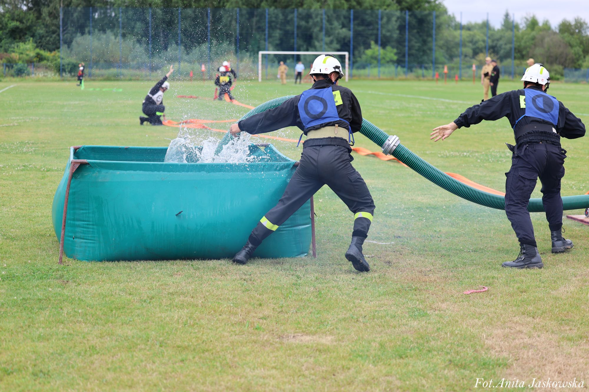 Dwóch strażaków w ciemnych mundurach i białych hełmach przy zielonym zbiorniku z wodą na trawiastym boisku sportowym. Woda rozpryskuje się z węża.