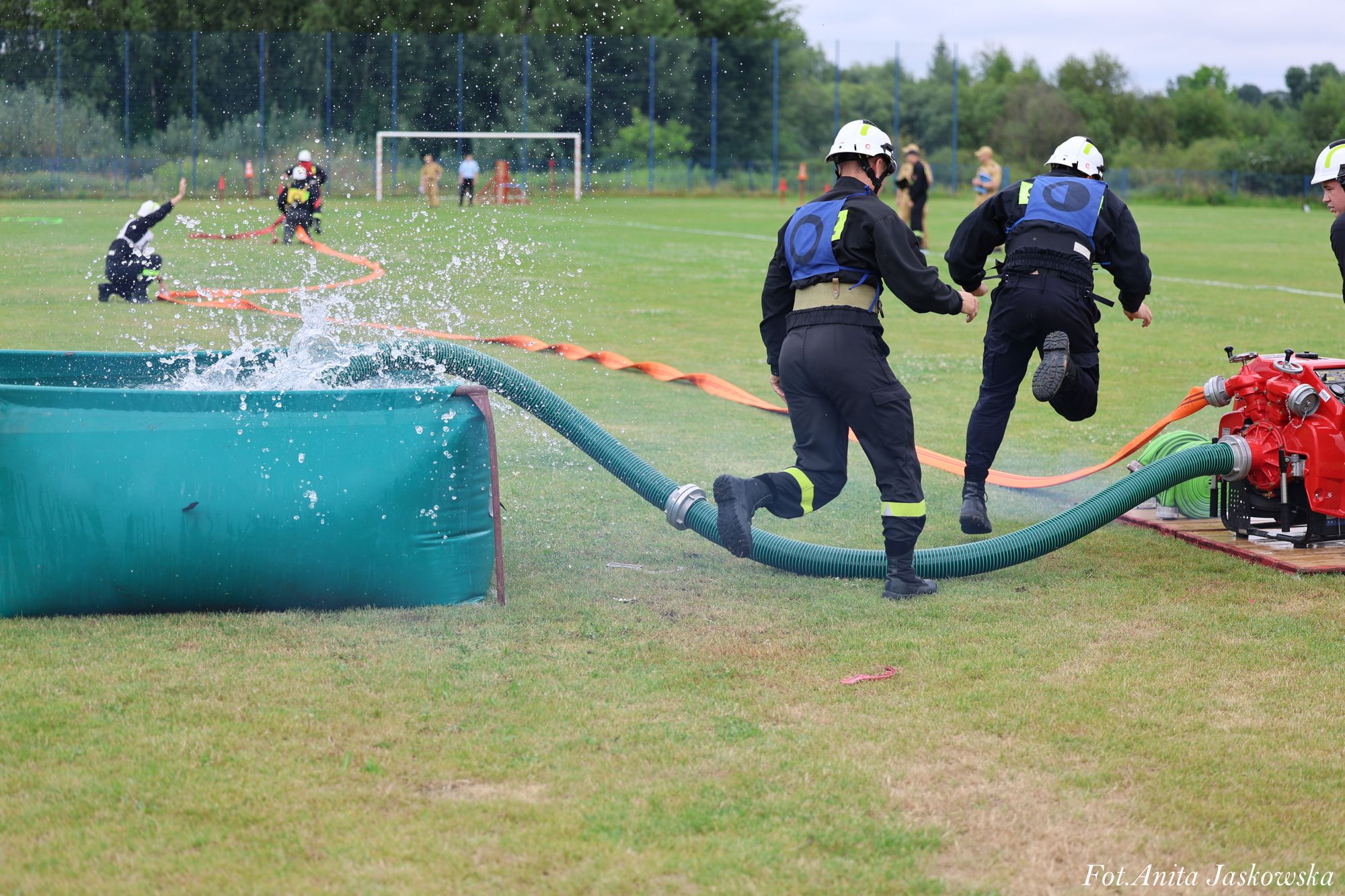 Dwóch strażaków w ciemnych mundurach i kaskach przy zielonym zbiorniku z wodą i czerwonym sprzęcie, w tle inni strażacy na zielonym boisku, krople wody w powietrzu.