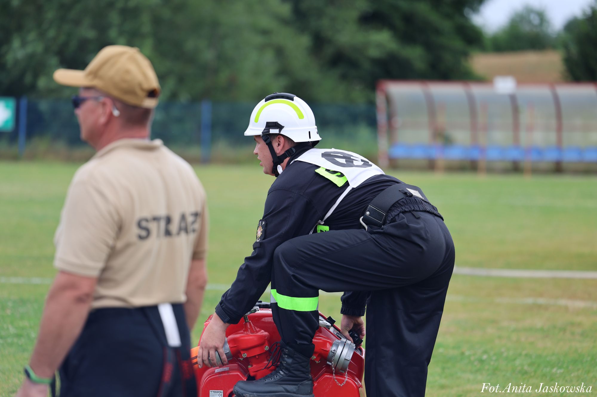 Mężczyzna w czarnym ubraniu strażackim i białym kasku pochylony nad czerwonym sprzętem na zielonym polu, obok drugi mężczyzna w beżowej koszulce z napisem STRAŻ.