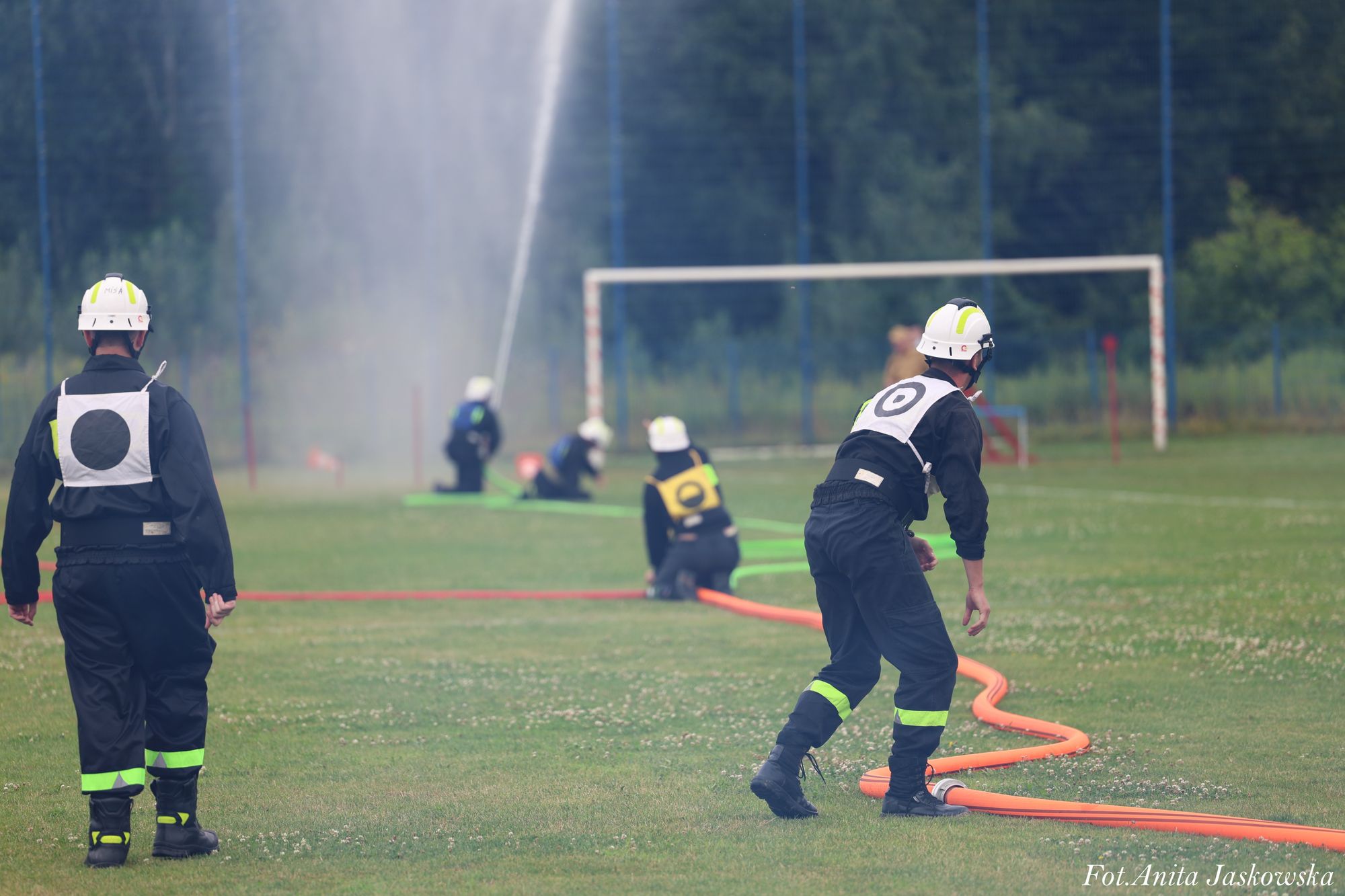 Strażacy w czarnych mundurach z odblaskowymi paskami i białych hełmach na zielonym boisku, rozkładają węże strażackie, w tle strumień wody z prądownicy.