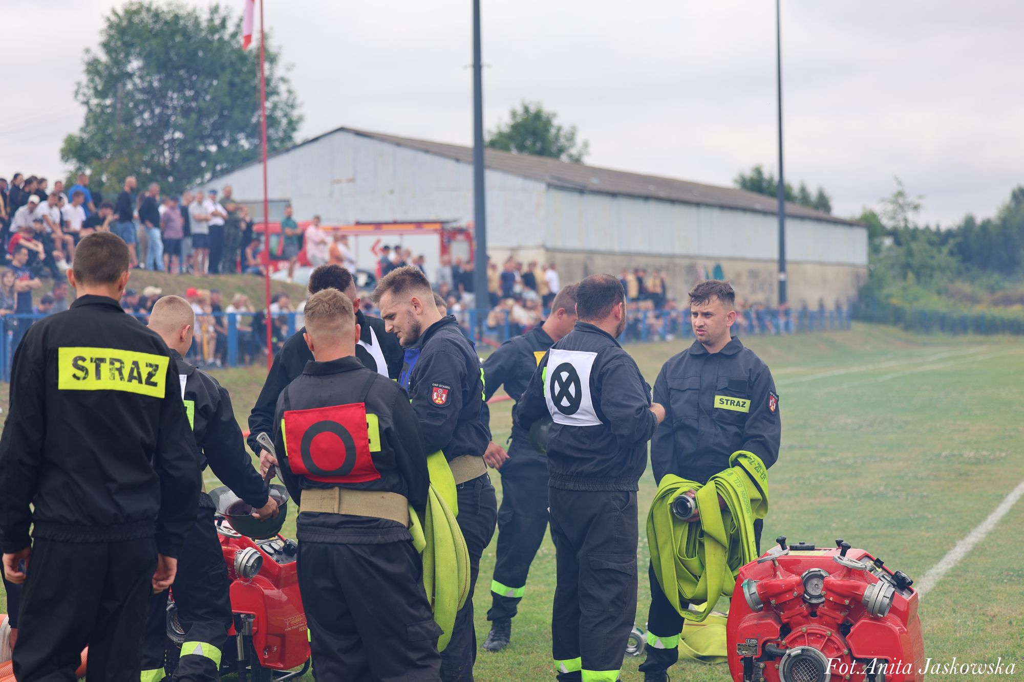 Grupa mężczyzn w czarnych mundurach z napisem "STRAŻ", trzymających zielone zwinięte węże strażackie, stojąca na trawie, w tle tłum ludzi i budynek.