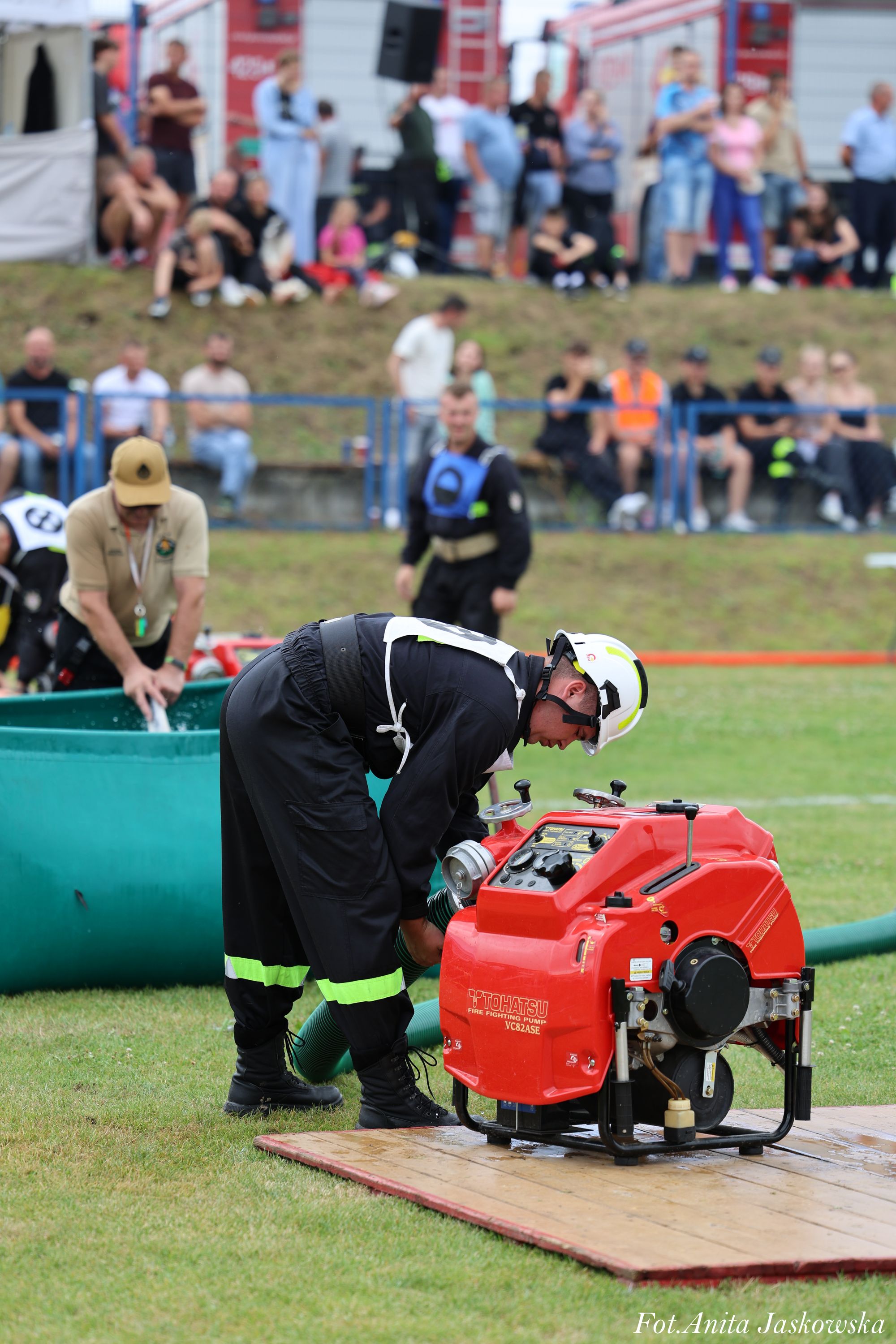 Strażak w czarnym ubraniu ochronnym i białym hełmie podłącza zielony wąż do czerwonej pompy pożarniczej na trawie, w tle ludzie i sprzęt gaśniczy.