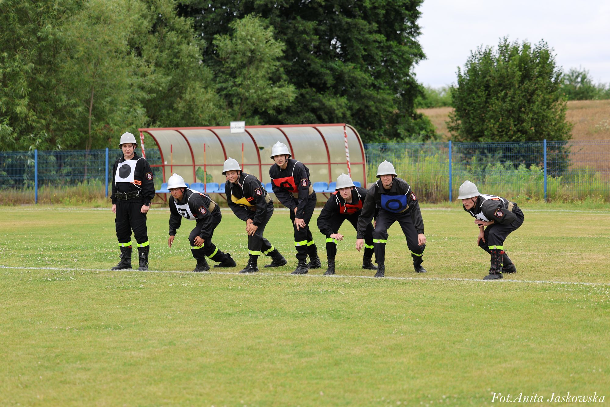 Siedem osób w hełmach i czarnych mundurach z kolorowymi oznaczeniami, pochylonych na zielonym boisku, w tle zadaszona ławka i drzewa.