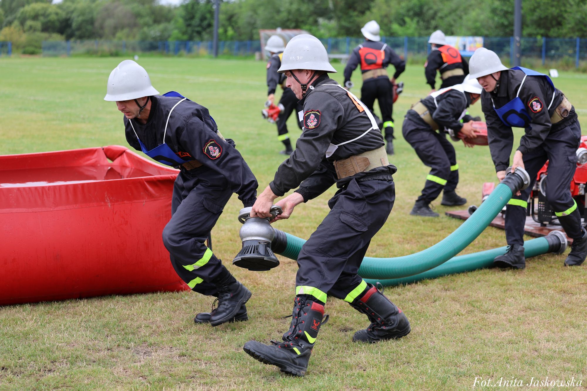 Strażacy w hełmach i czarnych mundurach z odblaskowymi elementami podłączają zielone węże do czerwonego zbiornika na trawie na otwartym terenie.
