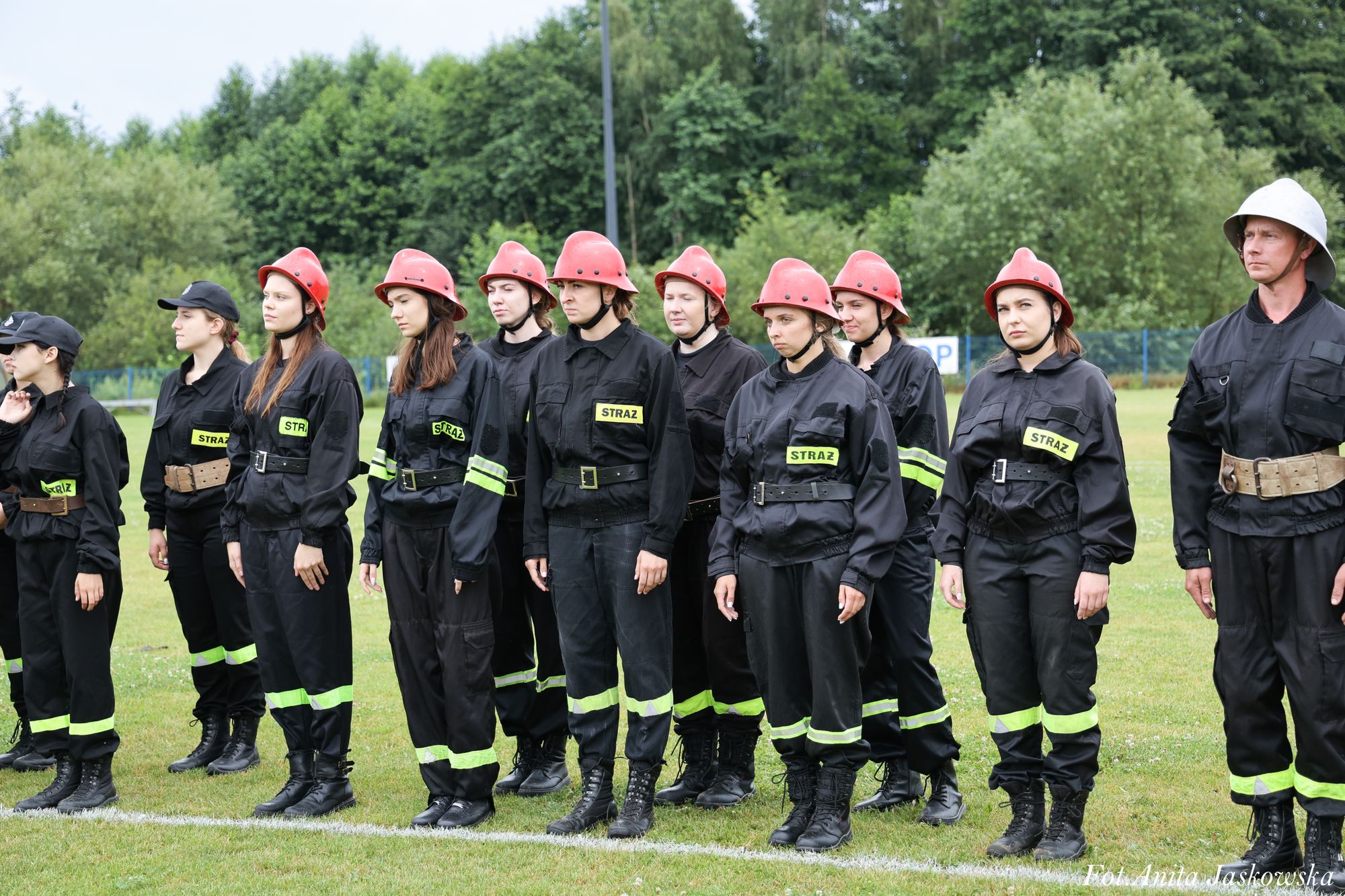 Grupa strażaków w czarnych mundurach z żółtymi odblaskami i czerwonych kaskach, stojących na trawiastym boisku na tle drzew.