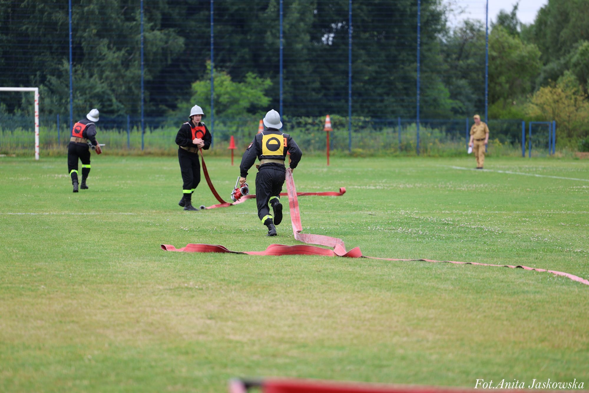 Trzech strażaków w czarnych mundurach z czerwonymi i żółtymi oznaczeniami, hełmy, rozwinięte czerwone węże na zielonym boisku sportowym.