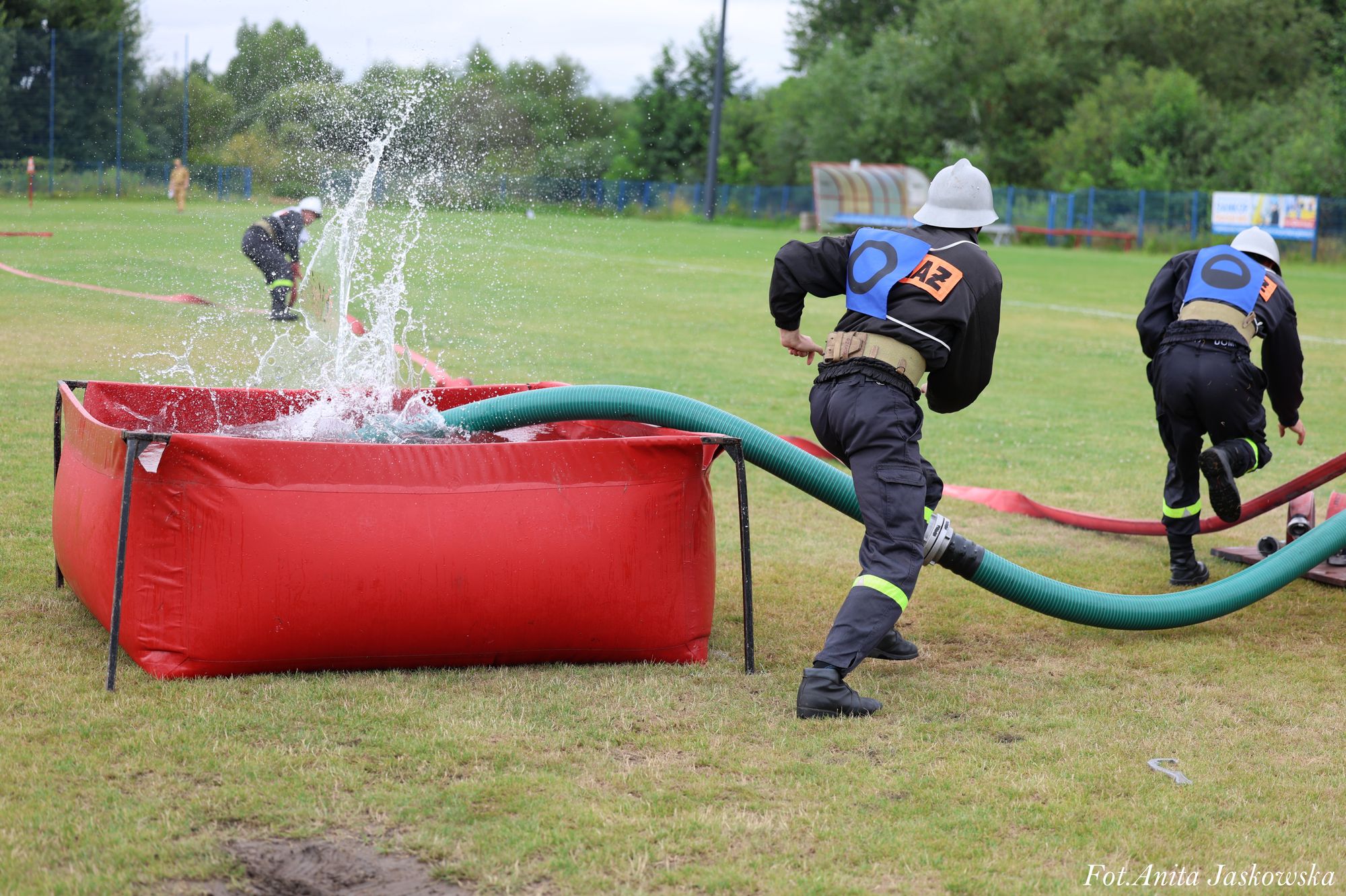 Czerwony zbiornik z wodą, zielony wąż, trzech strażaków w ciemnych mundurach i białych hełmach na zielonym trawniku, woda rozpryskująca się z węża.