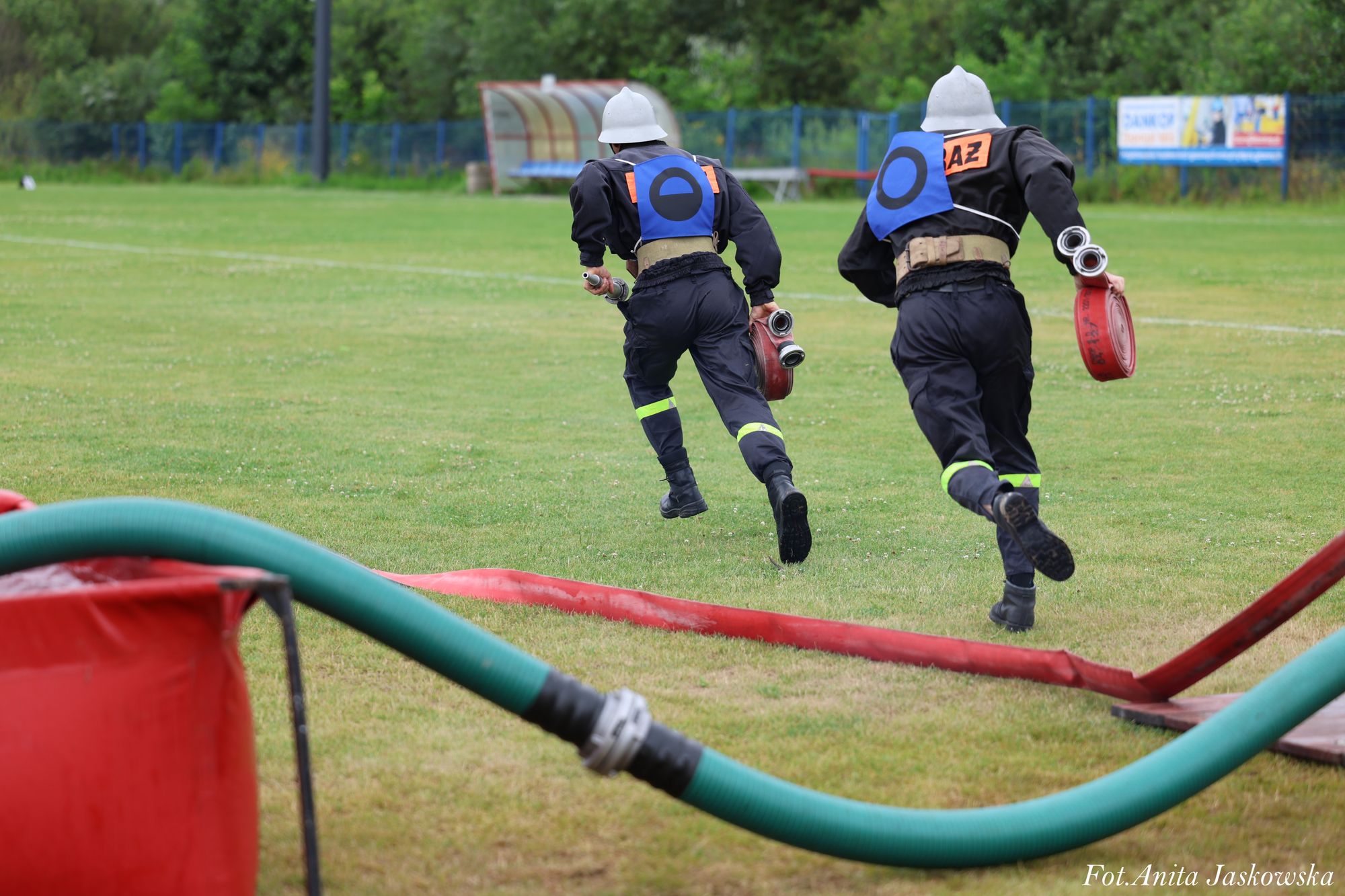 Dwóch strażaków w czarnych mundurach i białych hełmach biegnie po zielonym boisku, niosąc zwinięte czerwone węże strażackie, z niebieskimi oznaczeniami na plecach.