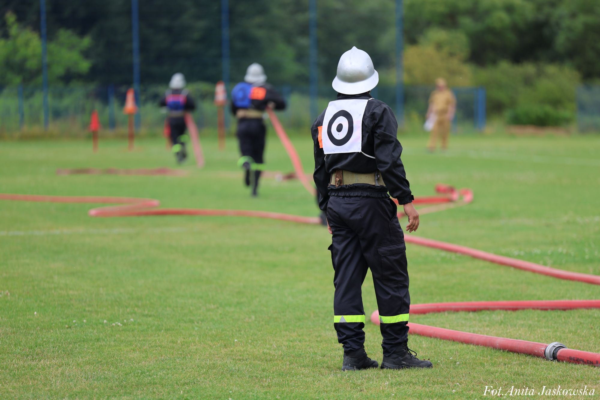 Osoba w czarnym ubraniu roboczym i białym hełmie stoi na trawie, na plecach ma biały panel z czarnym celem, w tle dwie osoby biegną z czerwonymi wężami strażackimi.