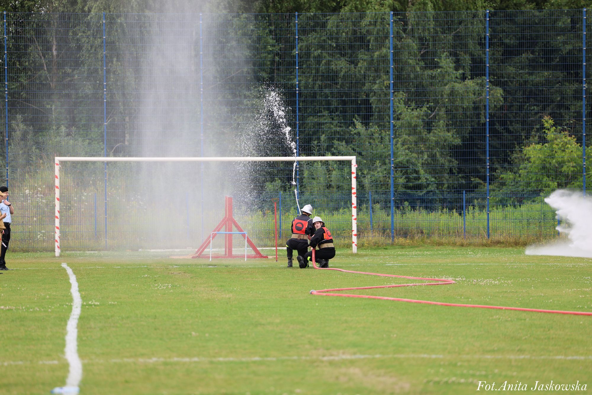Dwóch strażaków w hełmach i kamizelkach z numerami, trzymających wąż strażacki, polewających wodą na zielonym boisku sportowym, w tle niebieska siatka i drzewa.